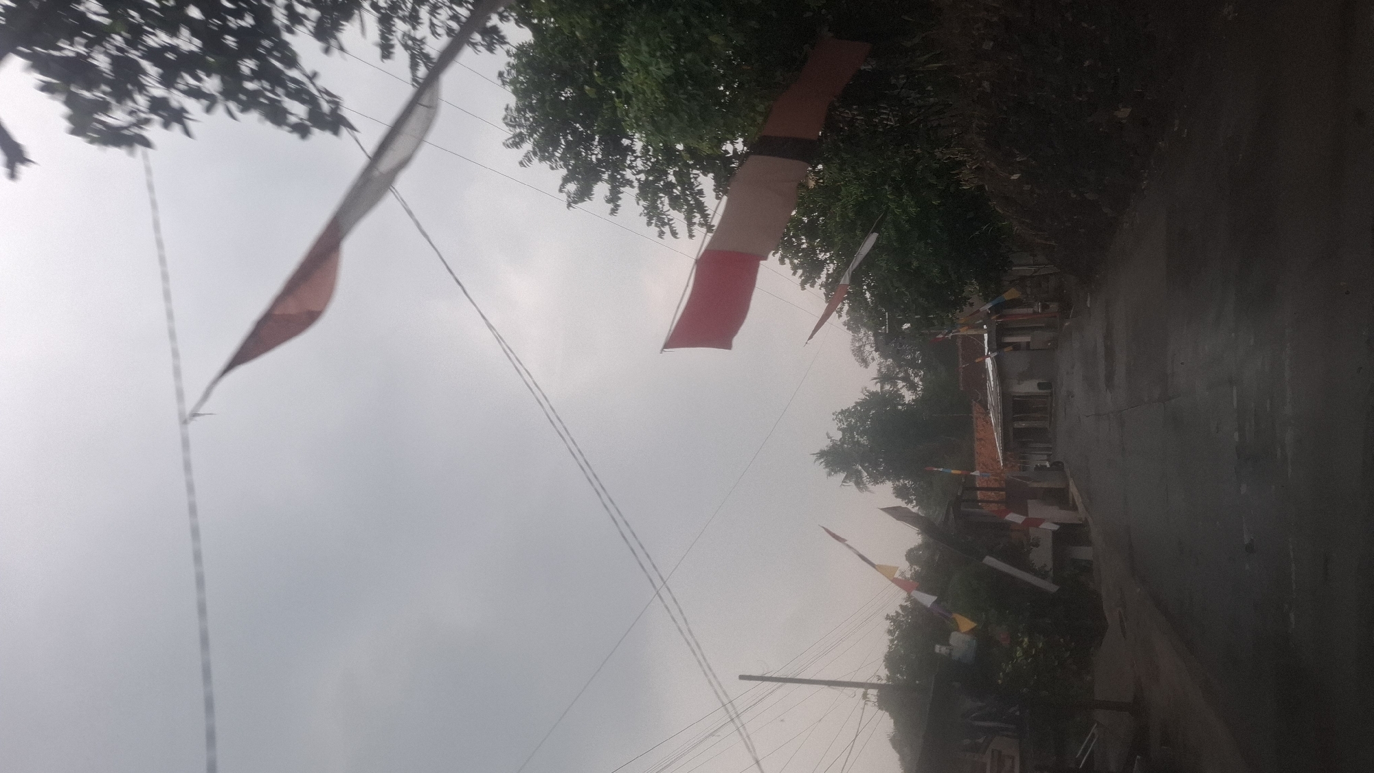 This image captures a street scene in Sirnagalih, Indonesia, under an overcast sky. A paved road runs through the center of the frame, leading towards a cluster of buildings. Several strings of red and white flags are strung across the road, fluttering in the breeze. These flags, known as "Panji Merah Putih," are a common sight in Indonesia, especially during national celebrations.
On the left side of the road, a simple building with a dark-colored roof and some visible window frames is present. Further down the street, a row of houses with red-tiled roofs can be seen. The buildings appear to be of modest construction, typical of residential areas in rural Indonesia.
Trees and foliage line both sides of the road, with some denser vegetation visible on the right, suggesting a somewhat hilly or uneven terrain. The sky is a uniform gray, indicating a cloudy or potentially rainy day. The overall impression is one of a quiet, everyday street in a community adorned for a festive occasion. No people are visible in the image, and the scene appears to be devoid of any immediate activity. The focus is on the environment and the decorative flags, highlighting the cultural significance of these symbols in the Indonesian context.