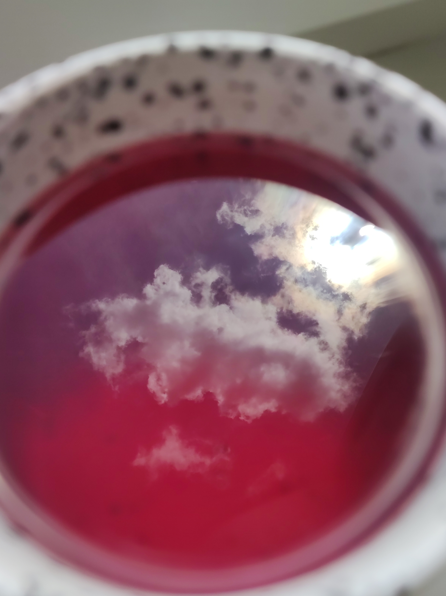 The image shows a close-up of a mug filled with a reddish liquid, likely a beverage. The surface of the liquid acts as a reflective mirror, capturing a reflection of the sky. The sky appears to be partly cloudy, with bright white and darker gray clouds illuminated by sunlight. The mug itself is white with small, dark speckled patterns around its rim. The lighting suggests it is daytime. The image does not contain any information to ascertain its location in Chaikovsky, Russia, or any specific details about people, actions, or interactions.