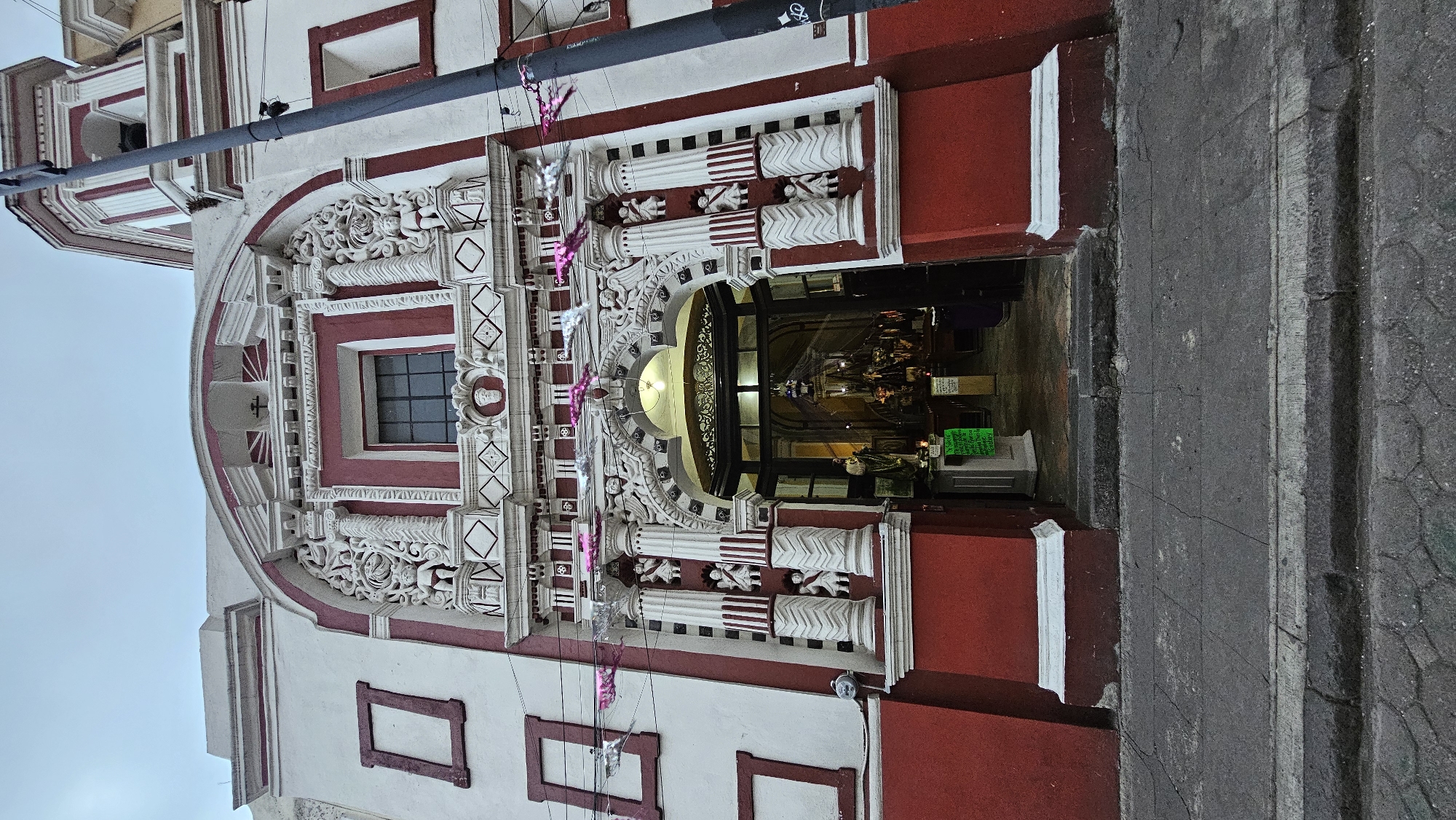 The image displays the intricately detailed facade of a building, characteristic of Baroque or Churrigueresque architecture often found in Puebla, Mexico. The dominant colors are off-white for the main structure and a deep reddish-brown or terracotta for accents, trim, and some architectural details.
The central feature is a grand arched entrance, framed by highly ornate white stonework carvings. These carvings include floral motifs, scrolls, geometric patterns, and figures, showcasing significant craftsmanship. Above the arch, a rectangular window with dark panes is set into a reddish-brown frame. Flanking the main entrance, pairs of columns with detailed bases and shafts are integrated into the facade, adorned with the reddish-brown banding and supporting decorative elements. Between some columns, small human figures or statues are visible.
Above the central window, the facade continues with more decorative elements, including a smaller arch and a relief featuring a cross within a sunburst design. Towards the top left (when oriented correctly), a portion of an upper story or bell tower is visible against a bright, overcast sky.
The entrance itself is a glass-fronted doorway, revealing a dimly lit interior. Reflections on the glass make it difficult to discern precise details inside, but internal lighting illuminates a light-colored counter or display, and a small green sign, the text on which is illegible.
Across the upper part of the facade, a thick black horizontal pipe or conduit runs. Hanging below this, festive decorations are strung across the entrance, consisting of shimmering silver and pink metallic shapes, possibly stars or ribbons, suggesting a celebratory period.
The ground level features a dark, textured sidewalk with visible cracks. The overall lighting suggests an overcast day or perhaps early evening. No people are clearly visible in the image.