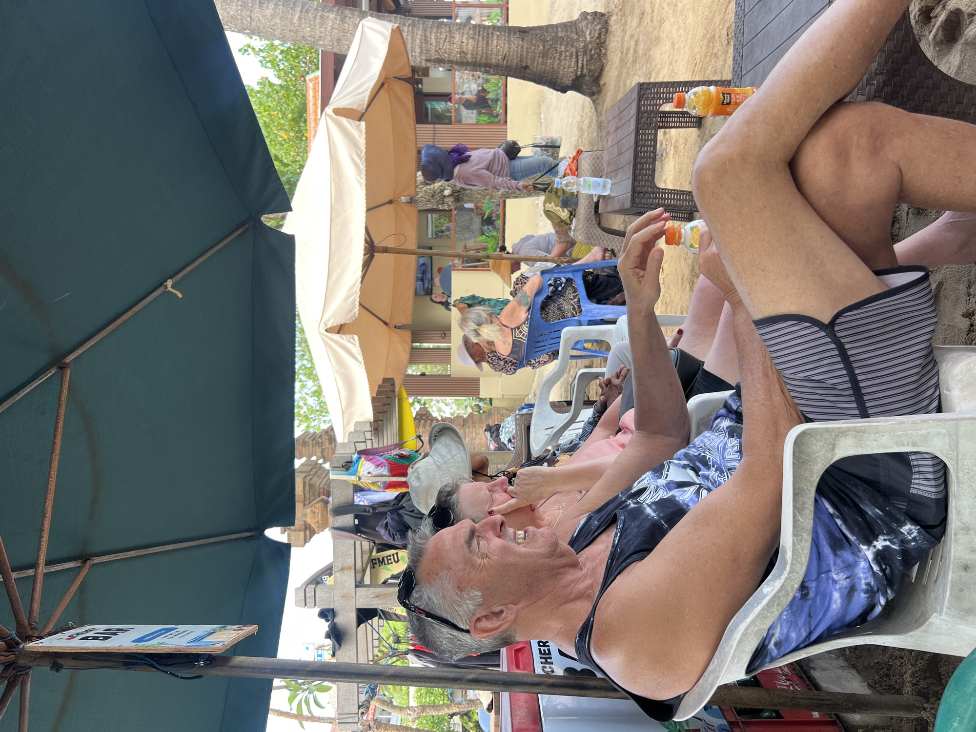 In Kuta, Indonesia, this photograph captures a candid scene under the shade of a large green umbrella and a beige beach umbrella. In the foreground, a man with grey hair, wearing sunglasses pushed up on his head and a black tank top with a blue and white tie-dye design, sits in a white plastic chair. He is laughing and looking to his left, with his arms resting on his lap. To his right, a woman, partially obscured, is also seated, wearing a pink top and a wide-brimmed hat. Her hand is raised to her face, and she is also looking to the left.
In the background, several other people are visible. A woman in a purple hat and sweater sits at a table in the sand, holding a bottle of water. Another group of people are seated further back, under the beige umbrella, some appearing to be tourists. Beach umbrellas and palm trees are prominent features of the environment, suggesting a tropical coastal setting. Bags and towels are scattered around, and there is a small structure with a sign that reads "BAR" hanging from the green umbrella. The sandy ground and the presence of beach chairs and umbrellas indicate this is likely a beachside cafe or bar.