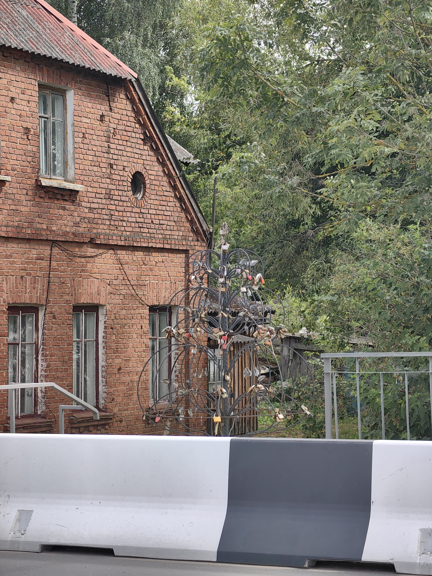 This image shows a street scene in Pastavy, Belarus, featuring a brick building with a red corrugated roof and several windows. In front of the building, a metal tree sculpture is adorned with numerous padlocks, a common symbol of love. A grey and white traffic barrier is positioned in the foreground, partially obscuring a metal railing and dense green foliage in the background. The weather appears overcast.