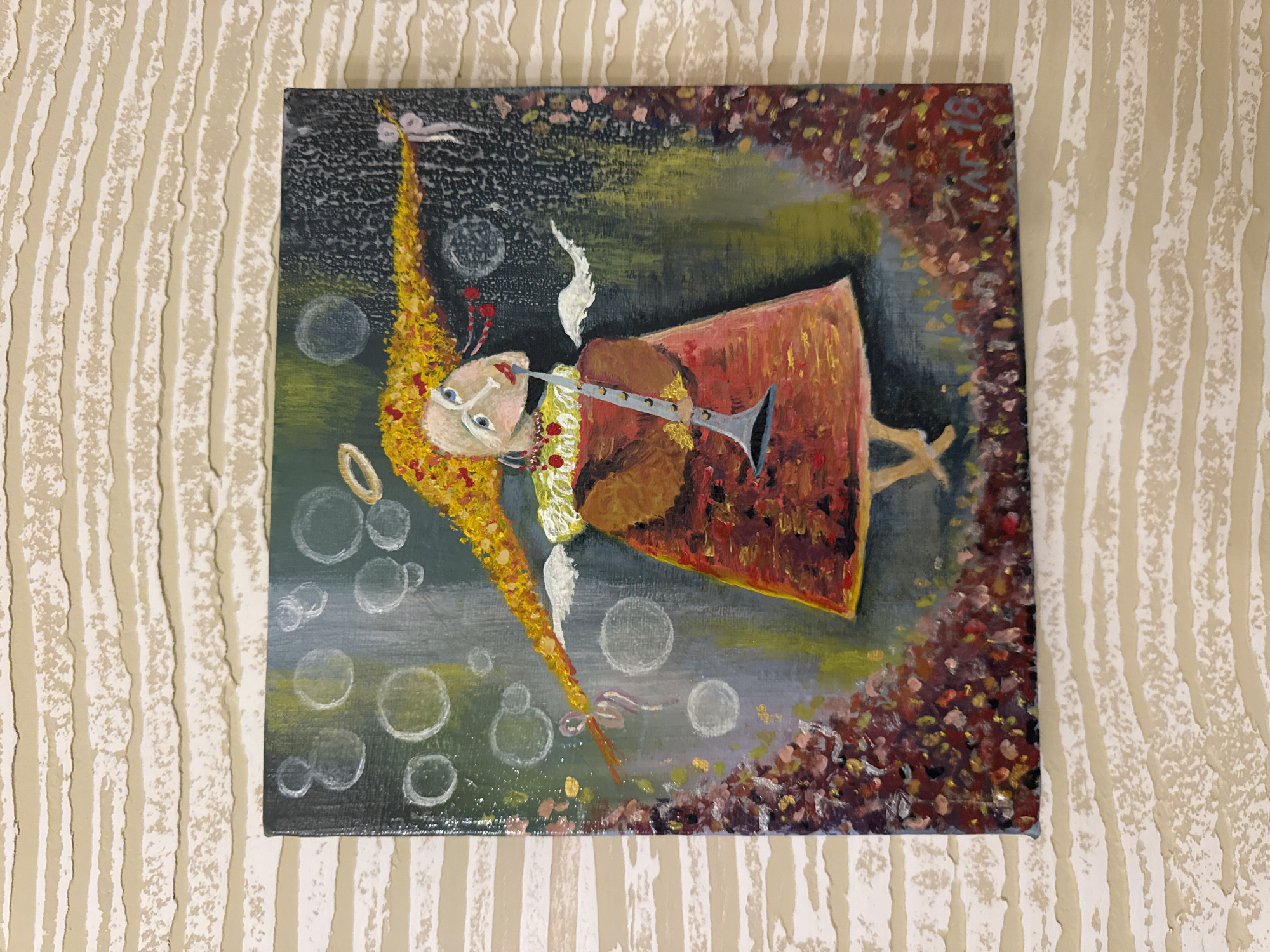 A painting depicts a stylized, childlike angel with a long, flowing yellow ponytail, tiny white wings, and a halo. The angel is wearing a reddish-orange dress and holding a clarinet to its lips, appearing to play it. The background features a dark, textured wash of greens and grays, with scattered white circles that could represent bubbles or light. The lower part of the painting is adorned with a dense, textured border of what appears to be red and brown floral elements. The artist's signature "AR 18" is visible in the bottom right corner. The artwork is hung on a wall with a textured, horizontal striped wallpaper. The painting is located in Yeysk, Russia.