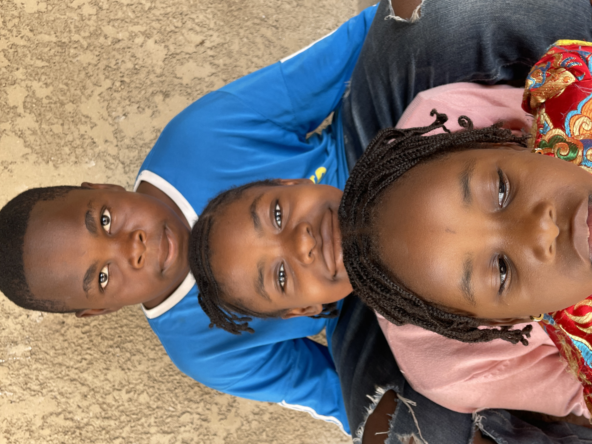 Three children are photographed from above. The child on the left wears a blue shirt with white trim. The middle child also wears a blue shirt, partially covered by the jeans of an adult person sitting behind them. The child on the right wears a light pink shirt and is partially covered by a red, gold, and blue patterned fabric. The children appear to be posing for the photo. The location could not be determined based on the image.