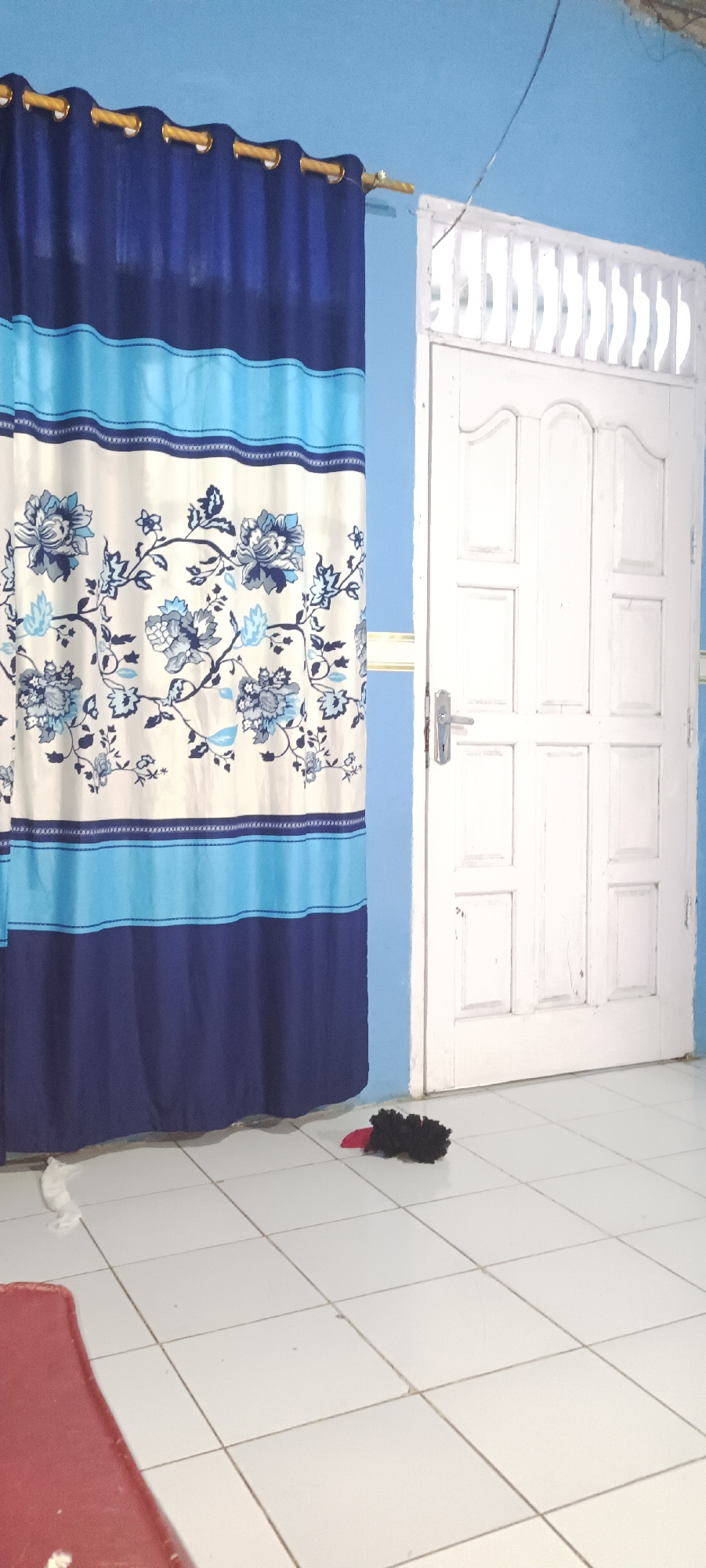 The image depicts an interior scene, likely a room, with a blue painted wall and white tiled floor. On the left side of the frame, a patterned curtain hangs, featuring floral designs in shades of blue and white, with horizontal bands of dark blue and light blue. To the right, a white wooden door with paneling and a silver handle is visible. In front of the door, on the floor, lies a small black object with a red accent, possibly a duster or a toy. A red rectangular object is partially visible in the lower left corner, potentially furniture. The overall impression is that of a simple, domestic space. This scene is located in Damuli Pekan, Indonesia.