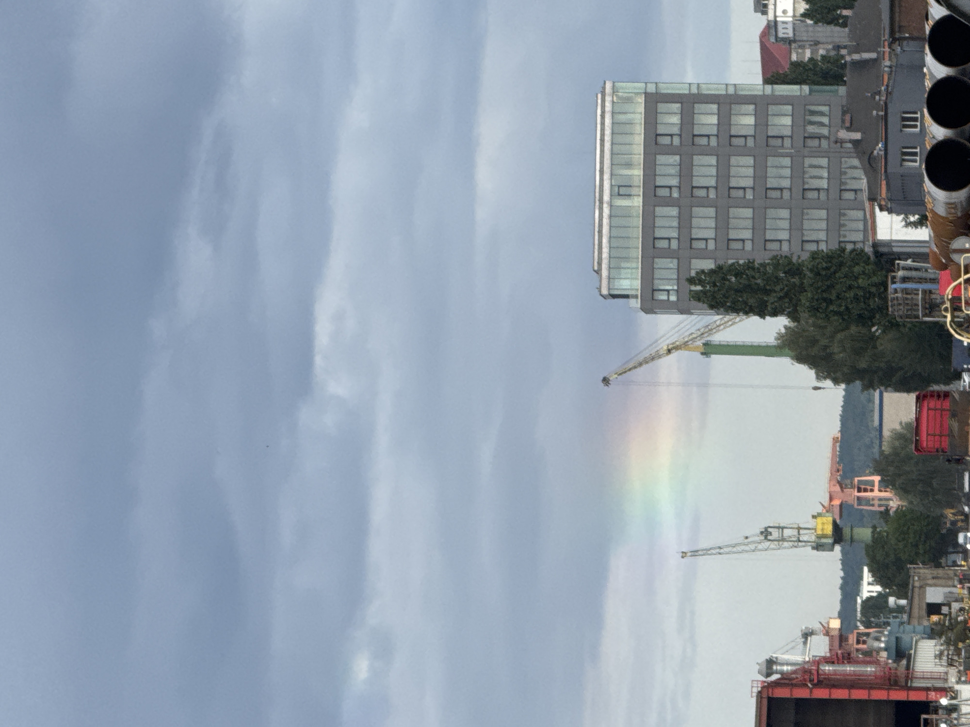 This is a medium shot, eye-level, outdoor photograph of a city scene with a cloudy sky. In the foreground, several large industrial pipes are visible, some made of rusted metal. Behind these, a red industrial building with a complex network of pipes and structures is situated on the left. To the right of this, a modern, multi-story office building with a glass facade stands prominently. Several construction cranes are scattered throughout the midground, some with yellow accents. A faint, partial rainbow is visible in the sky above the cranes. Trees are interspersed among the industrial structures and buildings. In the background, a wooded hill rises behind the city structures. The overall impression is of an active industrial or port area under an overcast sky, with a touch of natural beauty from the rainbow. The scene appears to be located in the industrial district of Gdańsk, Poland, likely near the shipyard or port facilities. There are no people clearly visible in the frame.