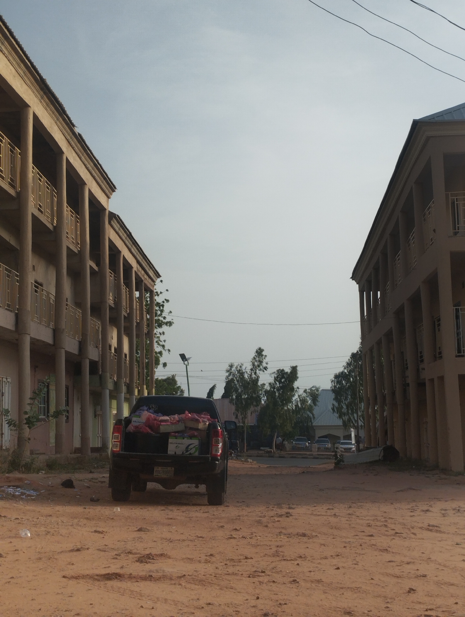 The image shows a dirt road in Gombe, Nigeria, lined with two-story buildings featuring open-air balconies with metal railings. A black pickup truck, loaded with items, is parked in the middle of the road, facing away from the viewer. In the distance, several parked cars and trees can be seen. Overhead, electrical wires stretch across the sky. The ground is dusty and uneven, with scattered debris.