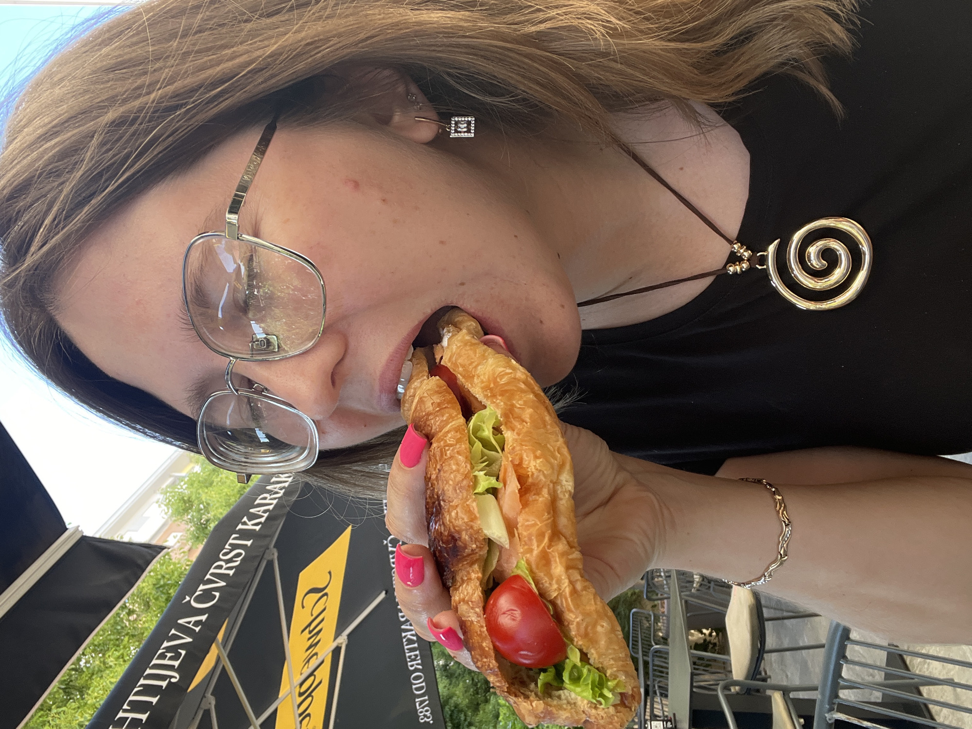 A person is depicted in a close-up, slightly overhead, and rotated view, taking a large bite out of a sandwich outdoors. The subject, a woman with long, light brown hair, wears modern, silver-rimmed glasses. Her visible ear features a small, square, silver-toned earring adorned with tiny clear stones. Around her neck is a brown cord necklace with a large, golden, spiral-shaped pendant. Her fingernails are painted a bright pink, and a delicate golden bracelet is visible on her wrist.
She is actively eating a sandwich made from a flaky, croissant-like bread, filled with fresh green lettuce and a prominent red cherry tomato, along with other lighter-colored fillings. The setting appears to be an outdoor cafe or restaurant in Podgorica, Montenegro, indicated by the visible metal outdoor furniture (chairs, table) and a black awning or umbrella. Bright sunlight suggests a daytime scene.
Visible text on the black awning/umbrella includes "HTIJEVA ČVRST KARAN" and "OD 1883," likely referencing a business name and its founding year. A yellow logo with stylized text, possibly "CUME," is also present. The overall image captures a casual moment of enjoyment while eating.