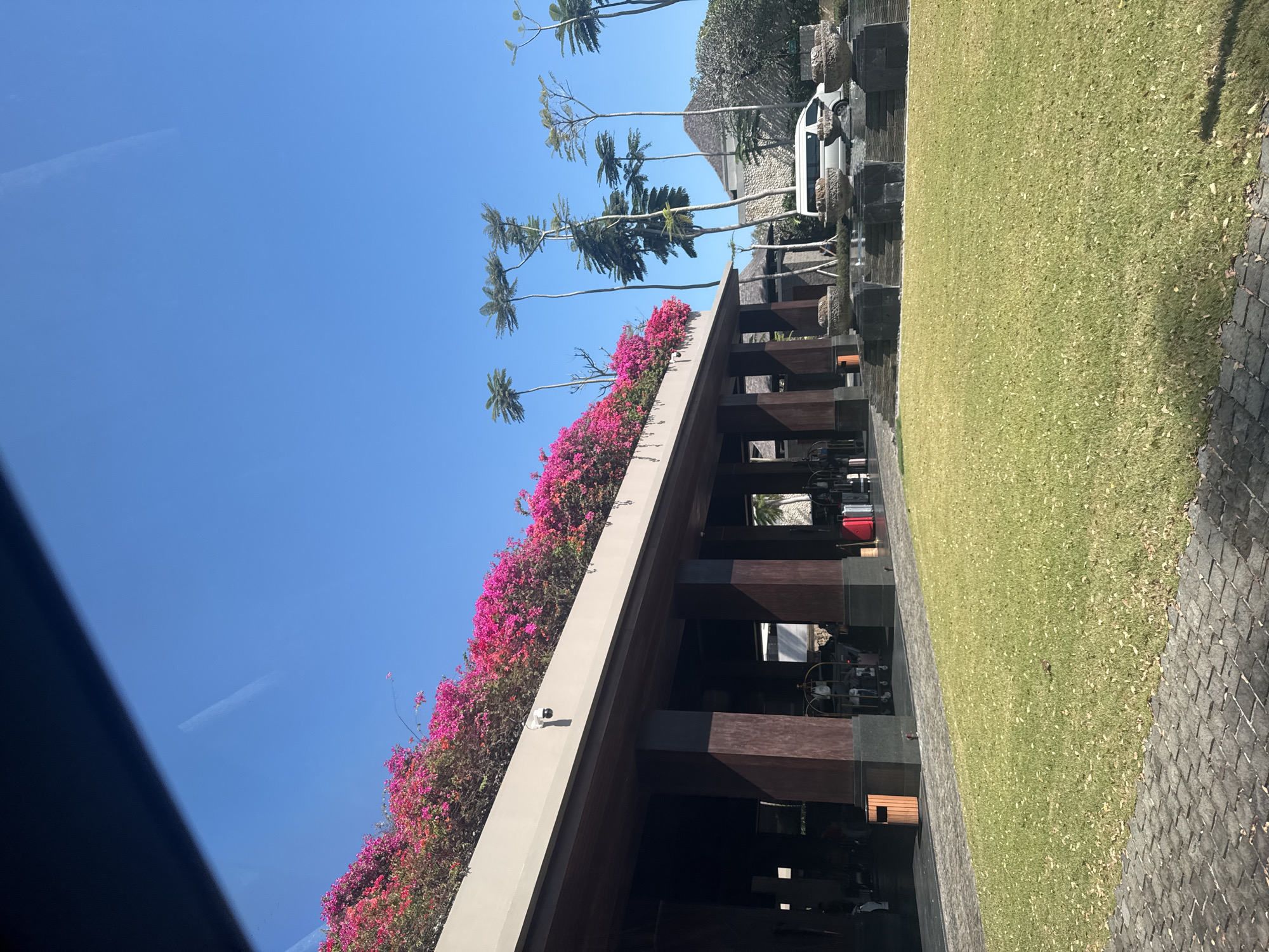 The image depicts the exterior of a modern resort in Kuta Selatan, Indonesia, bathed in bright sunlight under a clear blue sky. A prominent feature is the building's wide, covered entryway, supported by thick, dark wooden pillars. The roofline is adorned with a vibrant cascade of bright pink bougainvillea flowers, creating a striking contrast with the blue sky.
In front of the building, a well-maintained green lawn stretches out, scattered with a few fallen leaves. Paved walkways made of grey cobblestones run along the edges of the lawn and lead to the building's entrance. Several large, decorative stone planters are arranged in front of the building, adding to the landscaping.
Beyond the immediate foreground, a white van is parked on a paved area. In the background, there are glimpses of more tropical vegetation, including tall, slender palm trees with distinctive feathery fronds. A pyramid-shaped stone structure is also visible, suggesting traditional Indonesian architectural influences within the resort complex.
Inside the covered entrance, several bell carts with luggage are visible, along with some luggage on the ground, including two red suitcases. People can be seen in the distance, presumably staff and guests of the resort. A CCTV camera is mounted on the building's exterior wall, facing outwards. The overall scene conveys a sense of a tropical, upscale tourist destination.