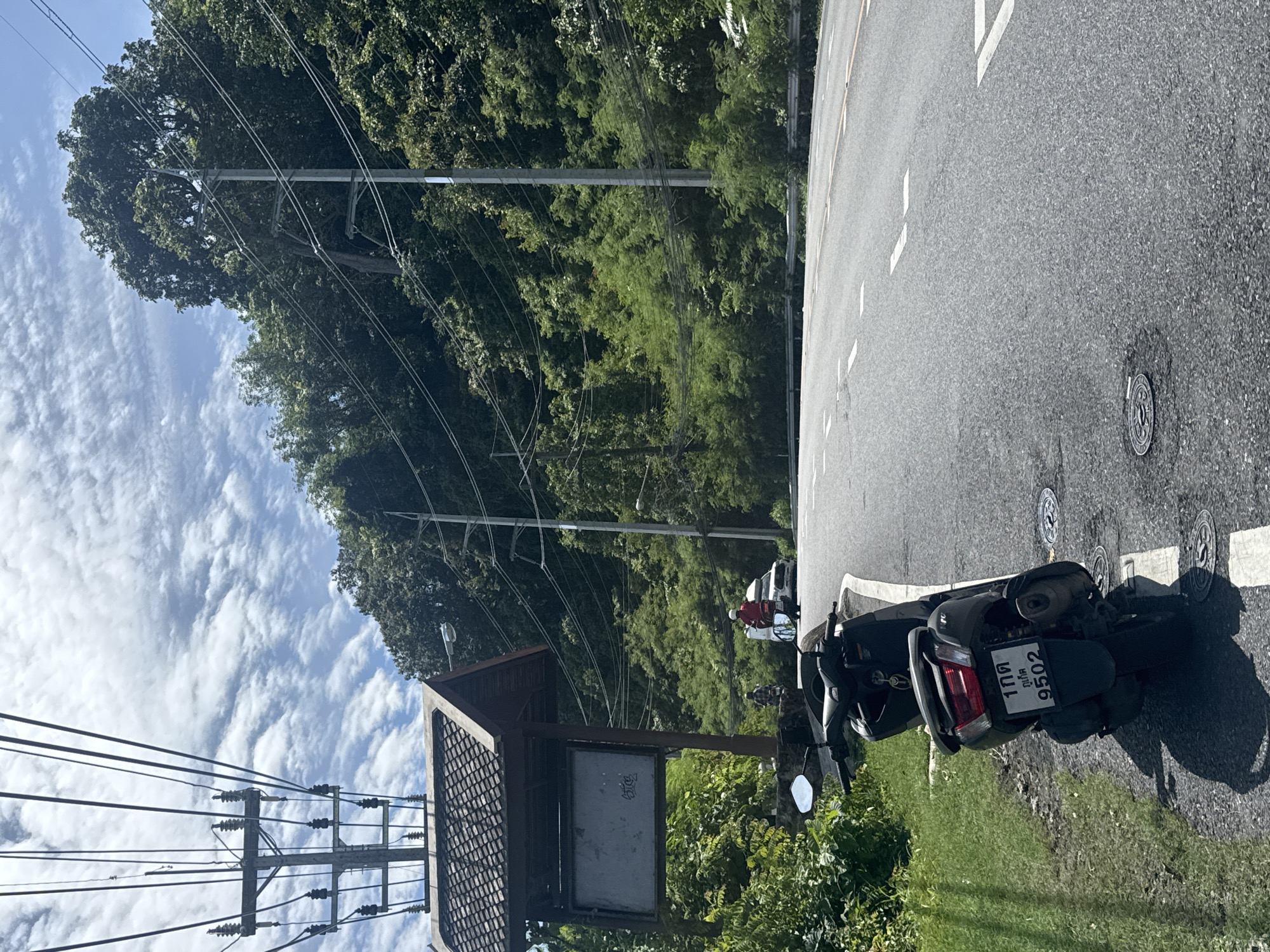 The image captures a scene on a paved road in Karon, Thailand. A dark-colored scooter is parked on the grassy shoulder to the left, with its license plate visible, displaying Thai characters and the number 9502. In the background, a man in a red shirt is riding a motorcycle, followed by a white car. The road curves to the right, bordered by a lush, green hillside covered in dense trees and vegetation. Overhead, a large utility pole with multiple wires crisscrosses the sky, which is partly cloudy with white, wispy clouds against a blue background. A small, tiled-roof structure, possibly a bus stop or information kiosk, is situated near the utility pole on the left. The overall environment appears to be a scenic, somewhat rural area with evidence of modern infrastructure.