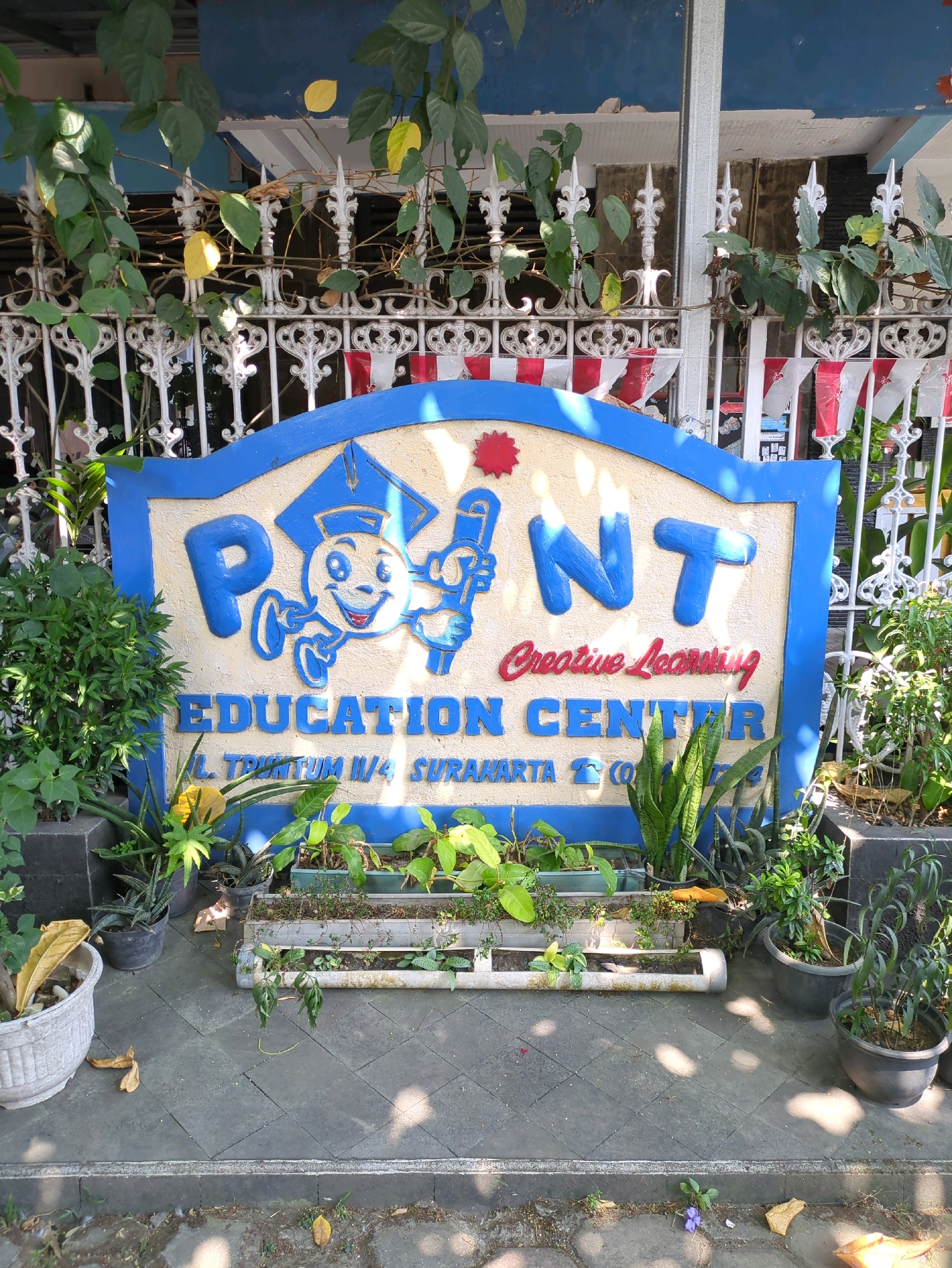 A low-angle exterior shot captures a sign for "POINT Creative Learning Education Center" in Surakarta, Indonesia. The sign, with its blue and yellow lettering and a cartoon mascot, is mounted on a textured cream-colored surface and framed by a blue border. Below the name, the address "JL. TRIKUTUM II/4 SURAKARTA" is visible. The sign is partially obscured by lush green foliage and plants in pots, creating a vibrant and natural foreground. A white metal fence with ornate decorative elements runs behind the sign, adorned with small red and white flags. Sunlight casts dappled shadows on the gray tiled ground. The overall scene suggests a welcoming educational environment situated within a tropical or subtropical climate, indicated by the abundance of greenery and the style of the building and fence.