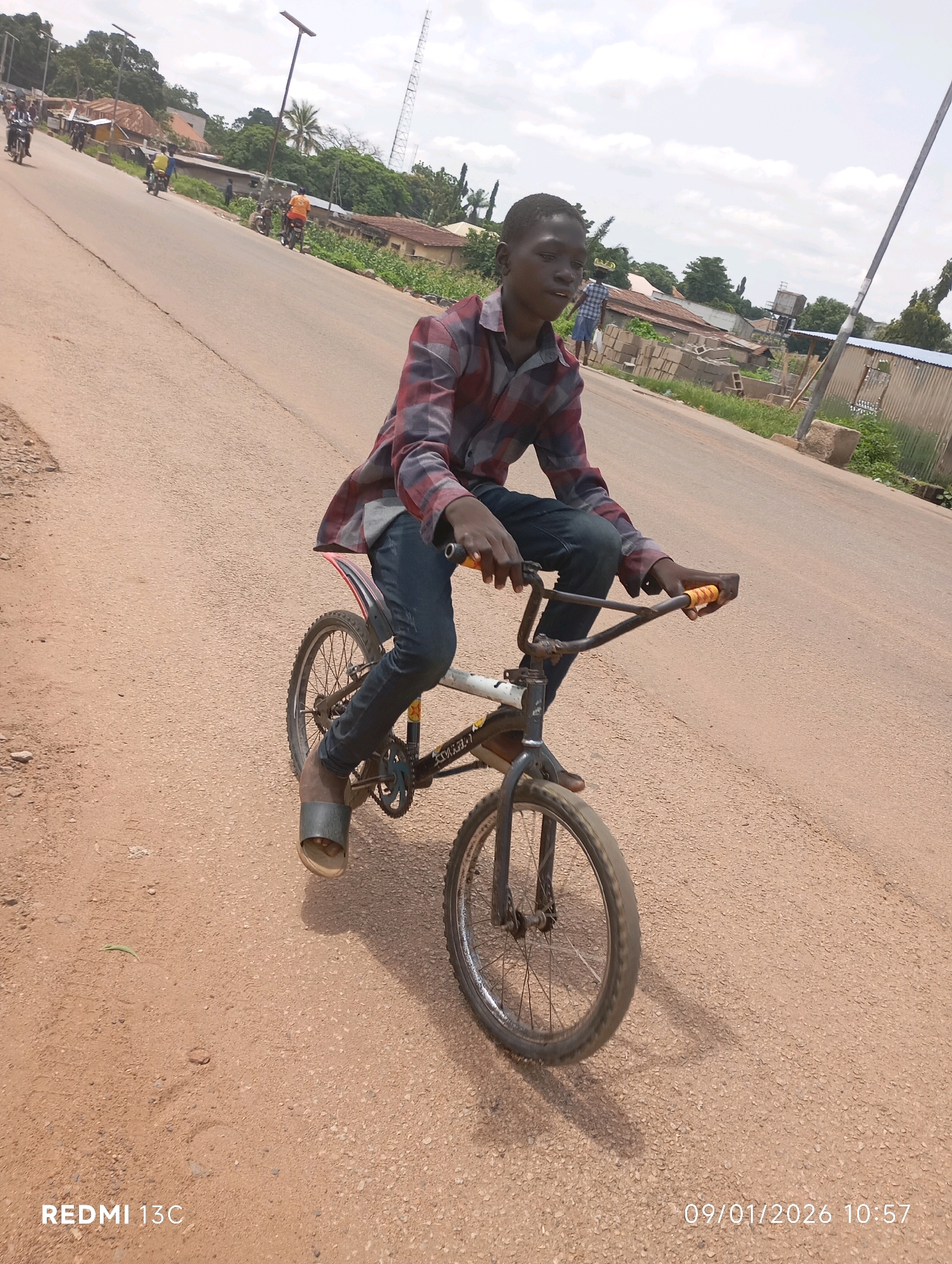 A young man in a red and black checkered shirt and dark blue jeans is riding a BMX bicycle on a paved road in Shendam, Nigeria. He is wearing dark colored sandals and his left foot is on the pedal. The bicycle is white and has yellow grips on the handlebars. The road is dusty and there are some loose rocks on the side. In the background, there are some buildings, trees, and a telephone pole. The sky is cloudy. The timestamp in the lower right corner reads "09/01/2026 10:57". The text "REDMI 13C" is in the lower left corner.