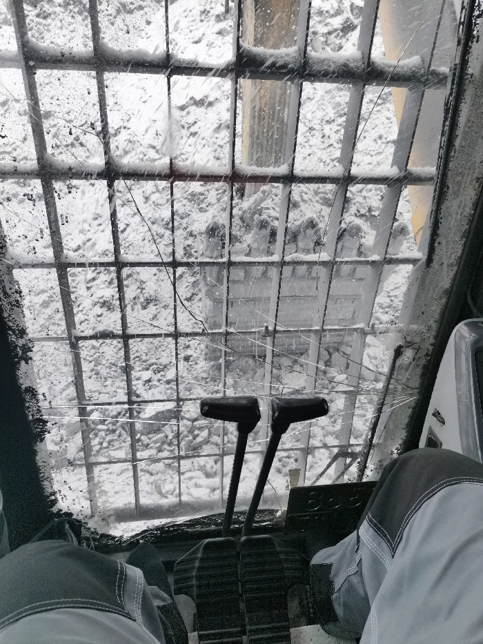 The image presents a first-person perspective from within the cabin of heavy machinery, looking outwards through a front window. The window is obscured by a metal protective grid, featuring numerous square openings, with fresh snow accumulated on its horizontal bars. The glass itself is dirty, streaked with ice or water, and exhibits multiple cracks, including a prominent diagonal fracture running from the upper-left towards the lower-right of the frame. Beyond the glass, the external environment is blanketed in snow, showing disturbed snow accumulation on the ground. A large yellow arm or boom structure, characteristic of construction or excavation equipment, is partially visible on the exterior, extending upwards on the right side of the view. Inside the cabin, in the lower third of the image, the legs of an individual are visible, clad in light grey work pants with darker grey knee sections. Two black foot pedals are present on the floor, and two vertical black control levers, each topped with a T-shaped handle, rise from the console immediately behind the pedals. The specific location within Im. Gastello, Russia, is not discernible from the visual elements in the image.