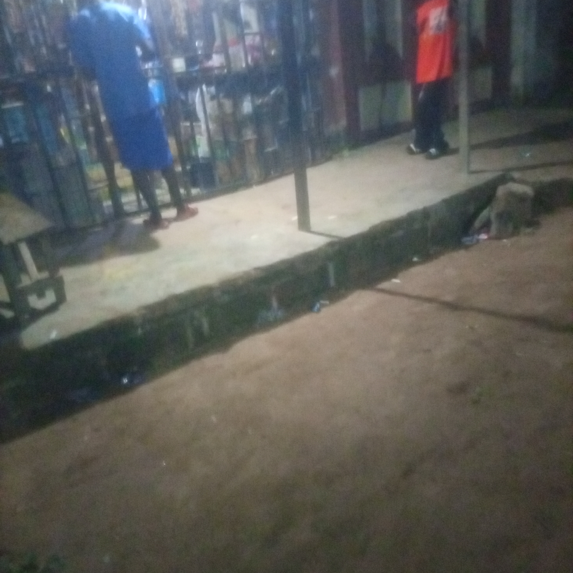 In an outdoor, dimly lit environment that appears to be a street or market stall, a person wearing a blue garment stands on a dirt path. To the right, another individual is visible, wearing an orange shirt and dark pants. The background consists of shelves or racks filled with various items, suggesting a commercial area. A low wall or curb runs horizontally in the foreground. The overall scene is dark and somewhat blurry, indicating it was likely taken at night.