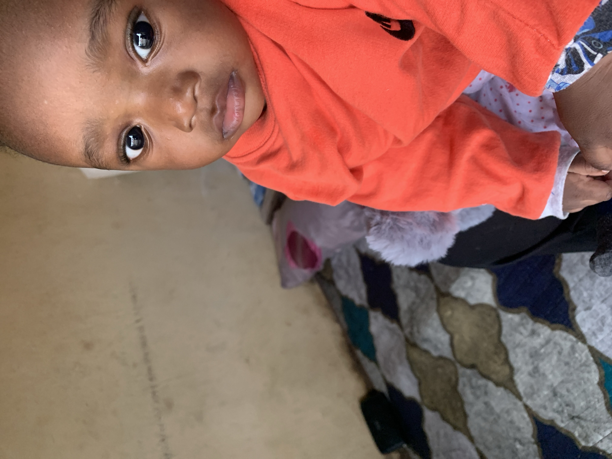 The image is a close-up, eye-level shot of a young child wearing an orange shirt and a white shirt with red polka dots. The child has dark skin and is looking directly at the camera with wide, dark eyes. Their mouth is slightly open. Behind the child, a patterned rug with blue, gray, and gold diamond shapes can be seen. A wall with a textured, light brown surface is in the background, with what appears to be faint markings or writing on it. A plush toy, possibly a stuffed animal, is partially visible behind the child, with its pink interior showing. The image appears to be taken indoors. The provided location of Jos, Nigeria, suggests an urban setting.
