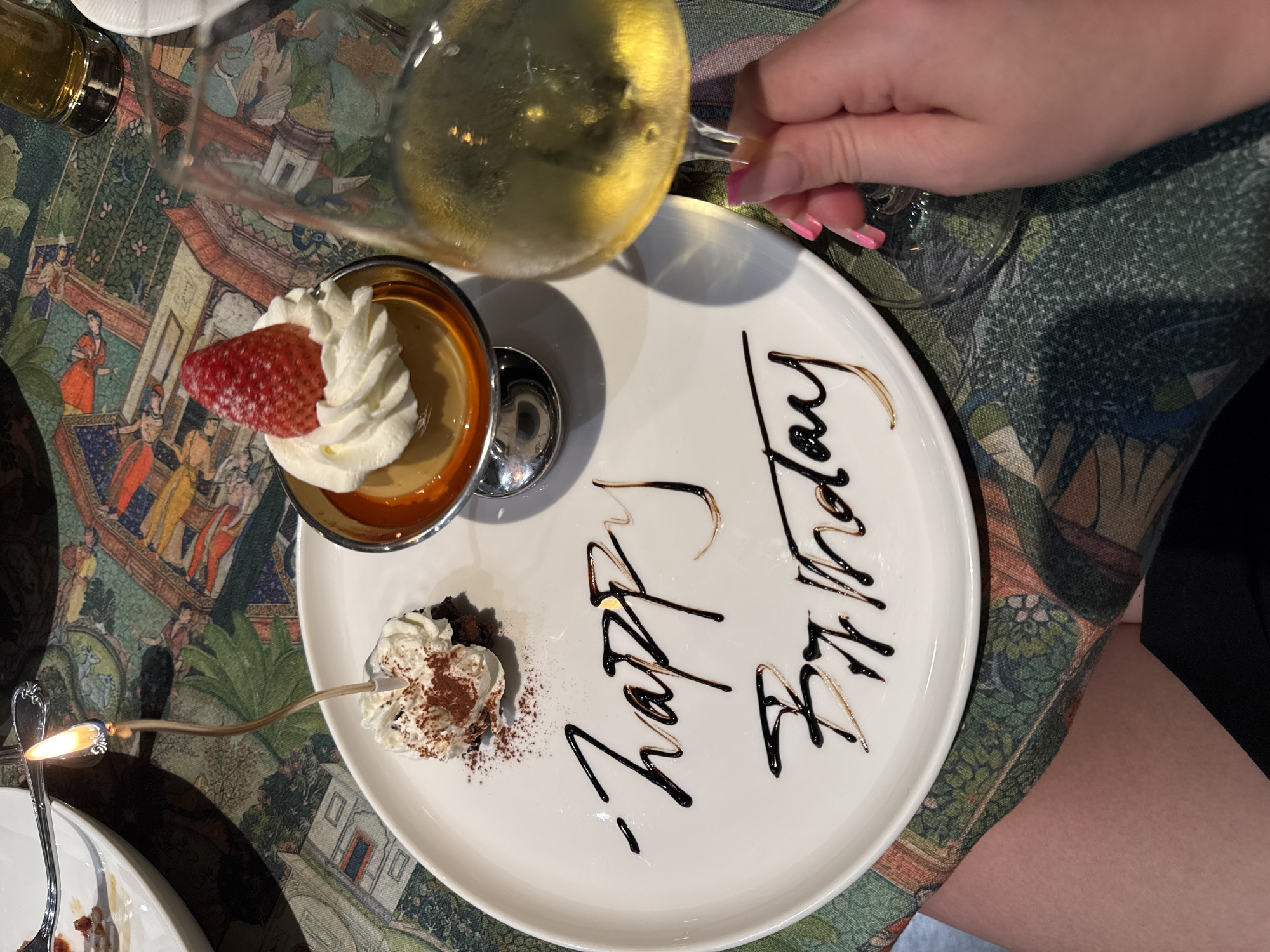 A dessert presentation is the focal point of a medium-angle shot, positioned on a patterned tablecloth and viewed from an elevated perspective. The dessert consists of a small ramekin filled with custard and topped with whipped cream and a strawberry slice, and a small mound of chocolate cake with whipped cream and cocoa powder. A silver spoon containing a lit birthday candle is placed near the chocolate cake. On the white plate, "Happy Birthday" is written in chocolate sauce. A hand holding a wine glass with a light-colored beverage is visible in the upper right corner, suggesting a toast. The setting appears to be a restaurant, indicated by the tablecloth and plate presentation. Zhangjiajie, China is not discernible from the details present in this image.