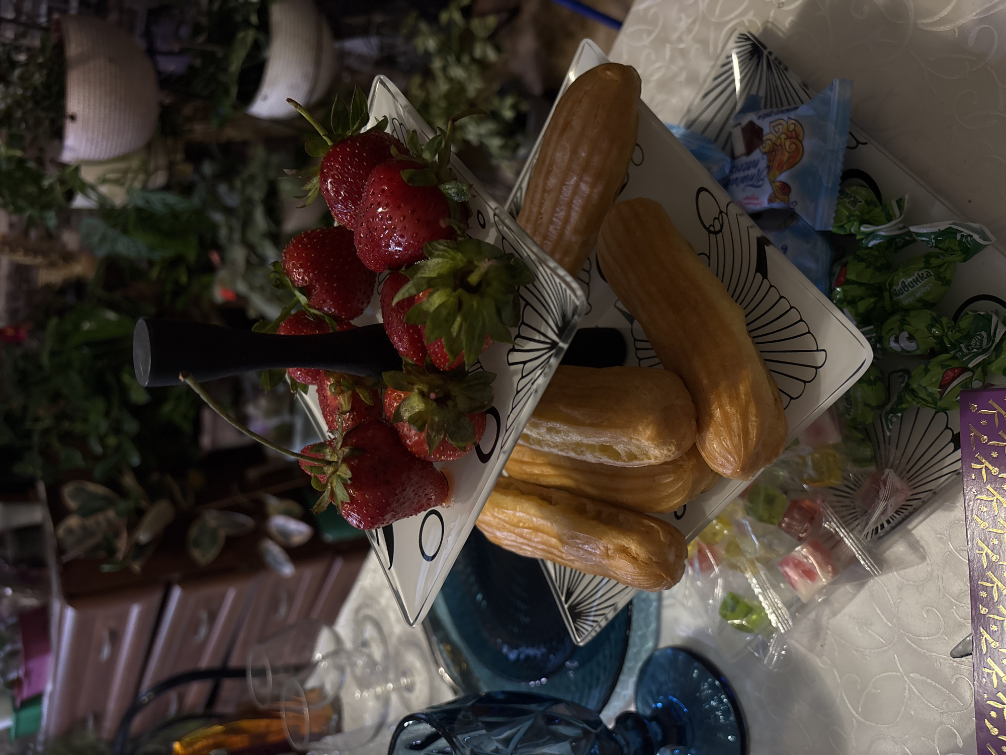 A tiered serving stand displays an assortment of desserts and fruits on a white tablecloth. The top tier holds several ripe strawberries. The middle tier is laden with éclairs, which appear to be filled with cream and topped with a glossy glaze. The bottom tier showcases a collection of individually wrapped candies, some of which are green and feature cartoonish designs, while others are in blue and light blue packaging. A blue glass goblet is visible to the left of the serving stand. The background suggests an indoor setting with wooden furniture and an abundance of potted plants, indicating a potentially domestic or restaurant environment. The image was taken in Sinyavino, Russia.