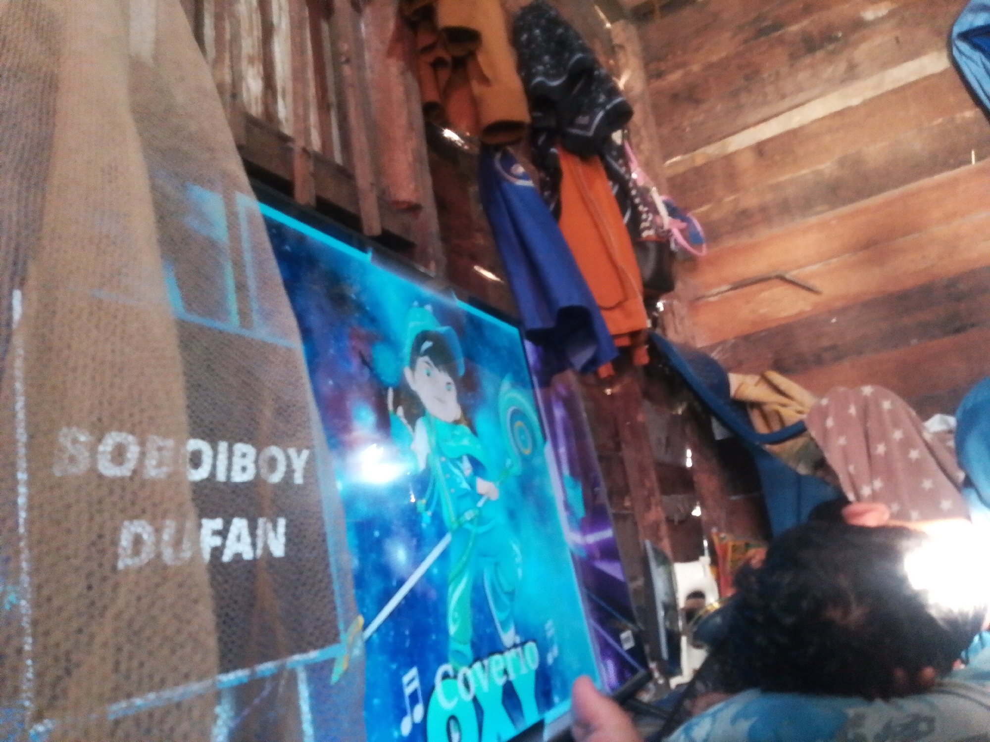 In Labuhan Bilik, Indonesia, a dimly lit room features a television screen displaying a cartoon character dressed in blue. On the left side of the image, text on a tan mesh material reads "SOBOY BOY DU FAN". Several pieces of clothing, including an orange garment and a blue cloth, are hanging from a wooden structure in the background. A person's head and shoulder are visible in the lower right corner, and another person is partially visible in the background on the left. The walls appear to be constructed of wooden planks.