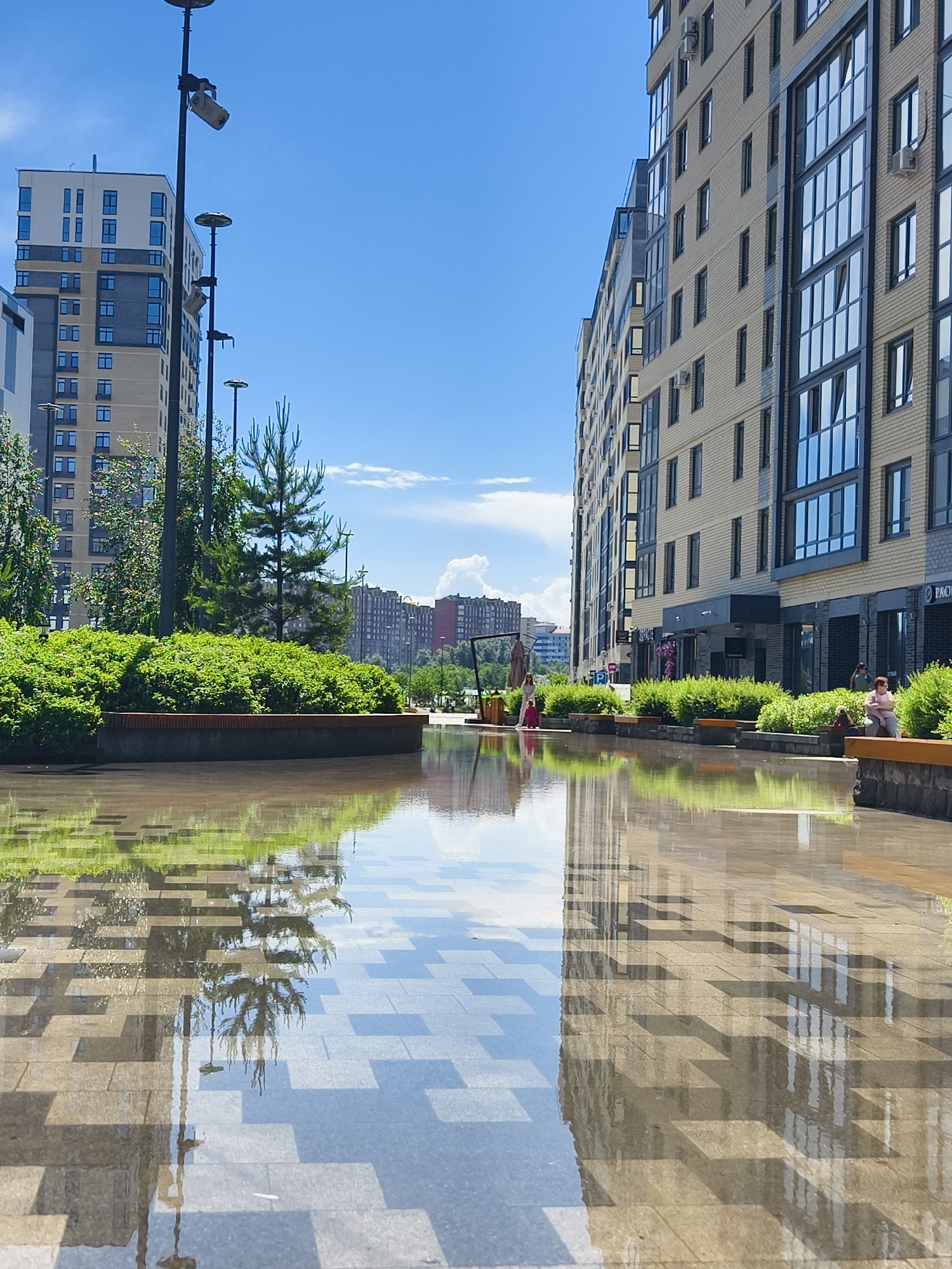 This image captures a vibrant urban street or plaza in Tyumen, Russia, under a clear blue sky with scattered white clouds. The setting features modern, multi-story apartment buildings flanking a wide paved area, which is partially covered by a thin layer of water, creating striking reflections of the sky, surrounding architecture, and greenery.
On the left, a tall, light-colored residential building with numerous windows rises into the sky. Its reflection is visible in the water. A row of contemporary streetlights with multiple fixtures extends along the left side, partially obscured by lush green bushes and small conifer trees planted in raised beds.
On the right, another modern apartment building with a cream-colored brick facade and large, dark-framed windows extends into the distance. Air conditioning units are visible on its exterior. Several individuals are present on this side of the plaza. One woman in a light top is seated on a wooden-topped bench integrated into a planter, looking towards the left. Further down, another person in pink attire is visible, and other figures are seen strolling or resting.
The water-covered paving in the foreground prominently reflects the surrounding scene, including the architectural patterns, the sky, and the trees. The patterns of the paving stones are also clearly visible beneath the water.
The scene depicts a peaceful daytime environment, suggesting a pleasant place for residents to relax and enjoy the outdoors. Visible text includes a parking sign with a 'P' and an arrow, a partially obscured sign on the right building that appears to start with "PAO" or "PAC", and a sign further down the street that reads "ЛЕГКО!" (LEGKO!). The overall impression is one of modern urban planning and leisure.