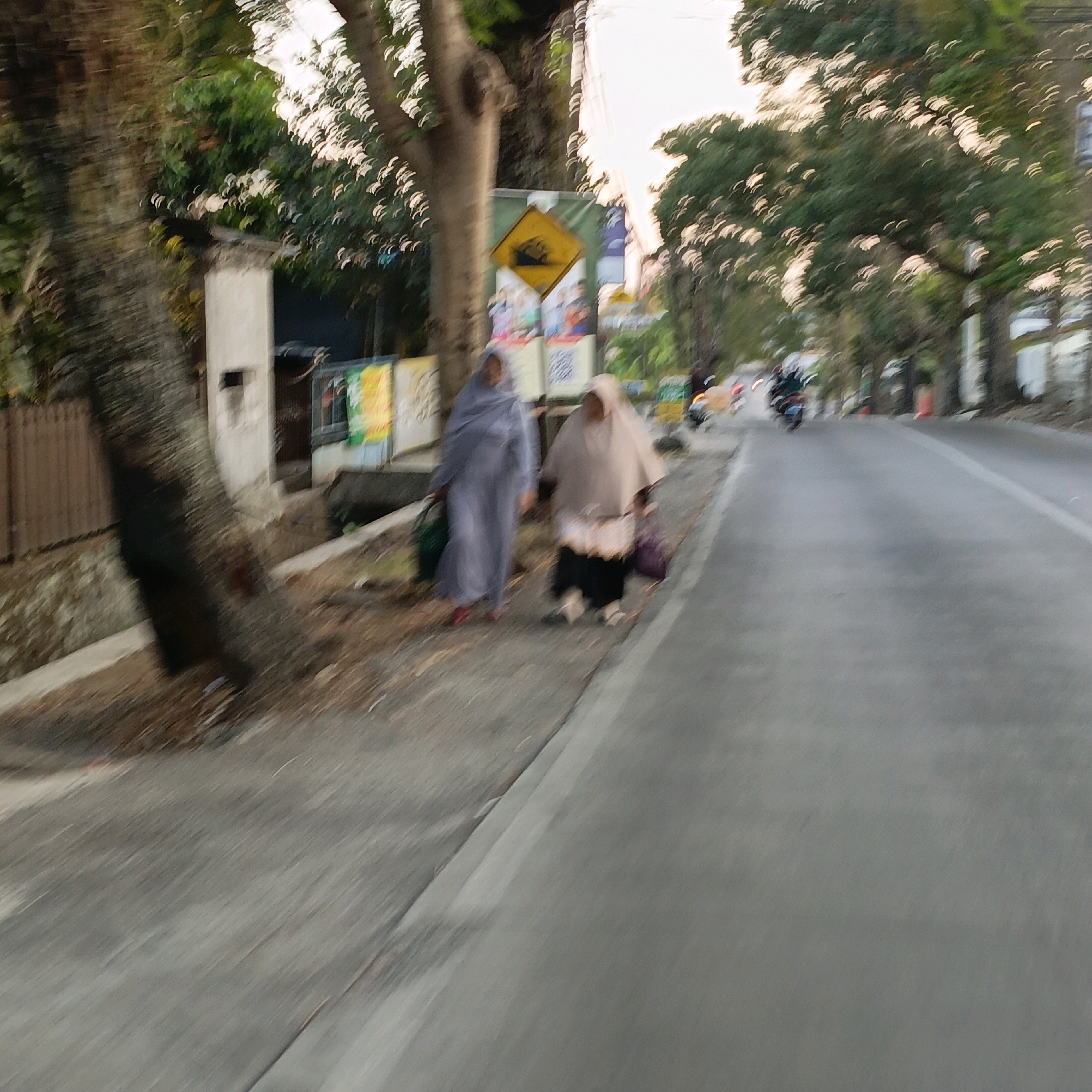 The image depicts a street scene in Salatiga, Indonesia, characterized by motion blur, suggesting a moving camera perspective. Two women, dressed in traditional Islamic attire, walk along a sidewalk adjacent to a road. The woman on the left wears a grey headscarf and a dark garment, carrying a green bag. The woman on the right wears a lighter-colored headscarf and a light-colored garment over a dark skirt. A yellow diamond-shaped traffic sign indicating a speed bump is visible on the sidewalk behind them. The environment includes trees lining the street, buildings in the background, and the road itself, with a white lane marker. Several motorcycles are also visible on the road, further contributing to the sense of movement. The overall lighting suggests it is either early morning or late afternoon.
