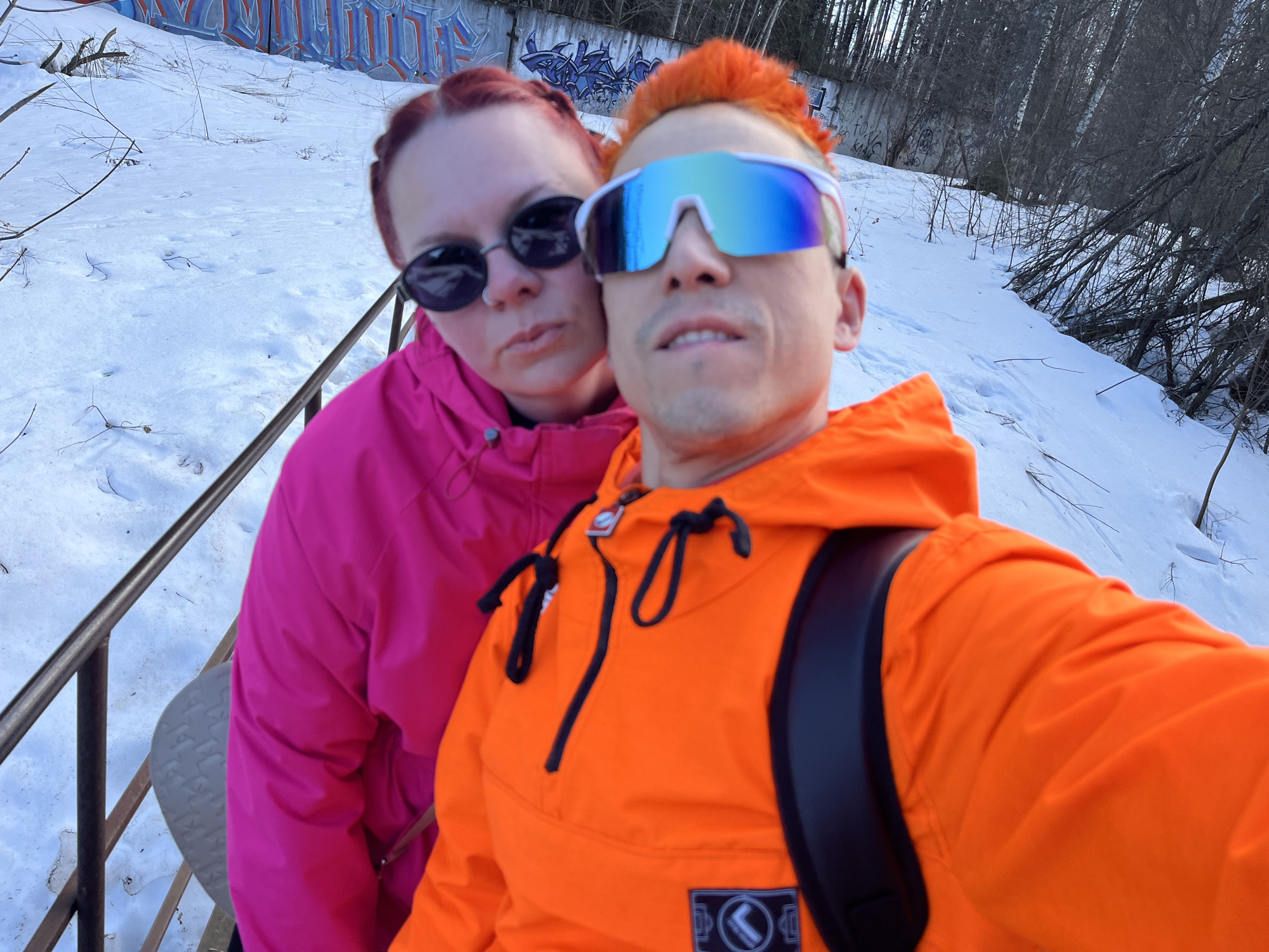 The image is a selfie of two people in a snowy, outdoor setting, likely in Chaikovsky, Russia. A person with orange hair and reflective sunglasses is in the foreground, wearing a bright orange hooded jacket and a black backpack. Behind them and slightly to the left is a person with red hair, wearing a pink jacket and sunglasses. The background includes a snowy slope with a metal railing on the left, and trees and graffiti-covered concrete in the distance. The weather appears cold and possibly overcast.