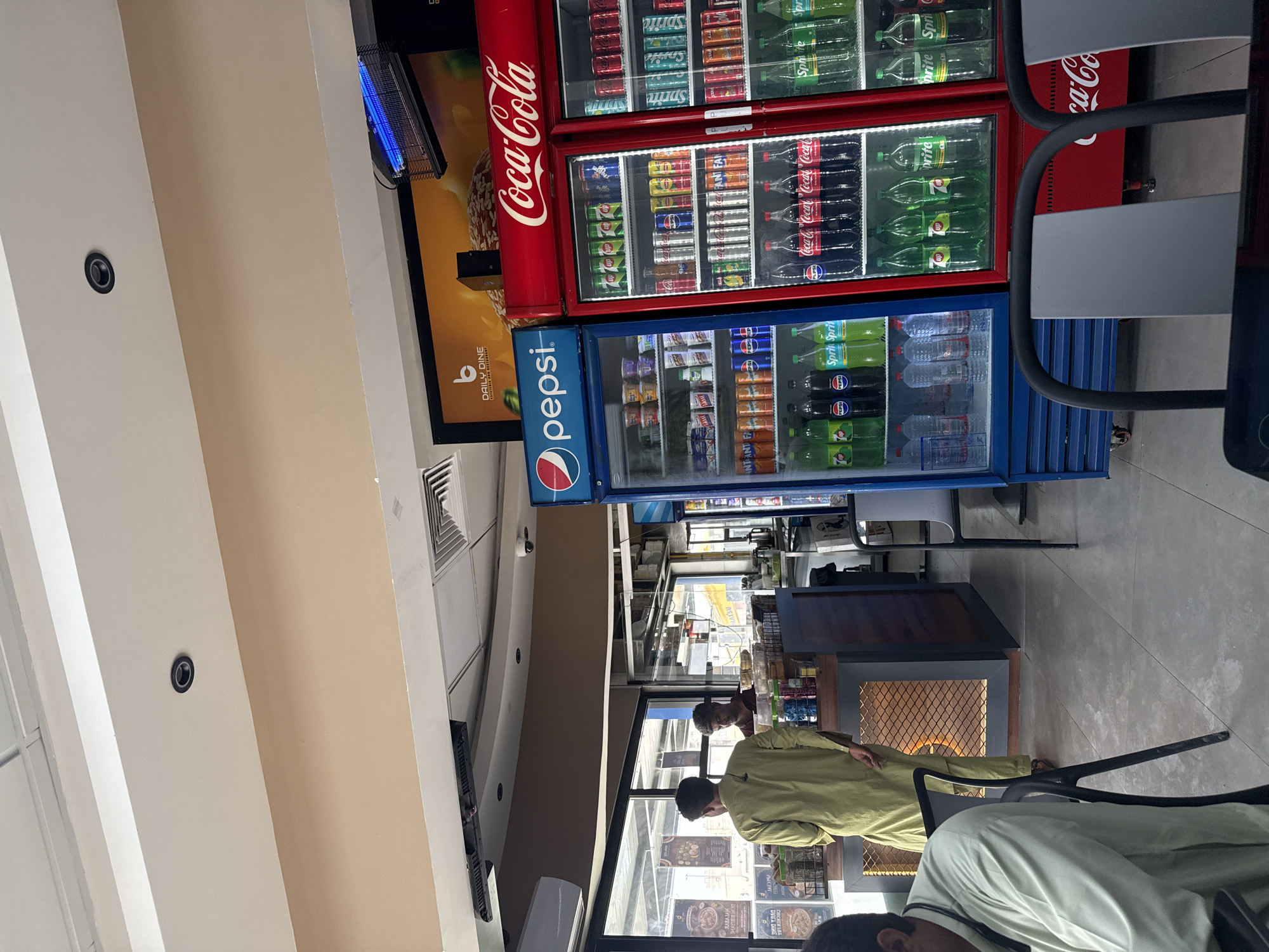 The image shows the interior of a restaurant or food establishment in Ras Al Khaimah, United Arab Emirates. In the foreground, a person in a light green traditional garment is standing near a counter. Behind the counter, another person is visible. The establishment features a display of beverages in refrigerators, including Pepsi and Coca-Cola branded drinks. Posters advertising food items are displayed on the wall behind the counter. The overall environment appears to be a casual dining or takeaway setting.