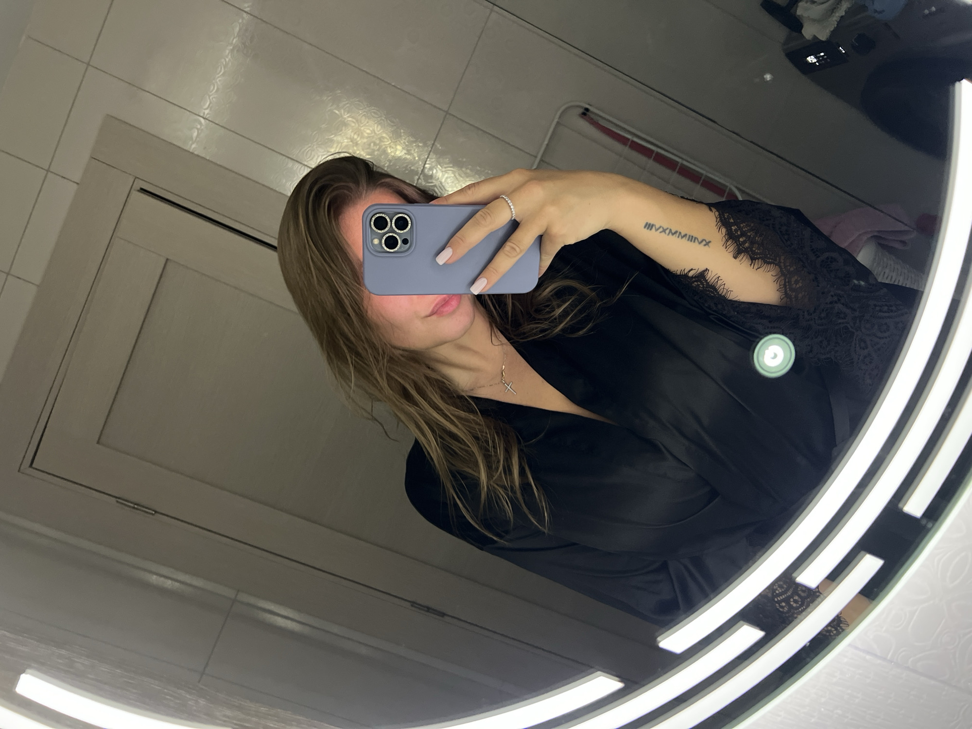 A woman in a black silk robe is taking a selfie in a bathroom in what seems to be an apartment or hotel. She is holding a light purple iPhone 13 with a ring on her finger. There is a tattoo in Roman numerals on her left forearm. The bathroom has white tile walls and a round mirror with LED lights. Based on the context, the location could be Budva, Montenegro.