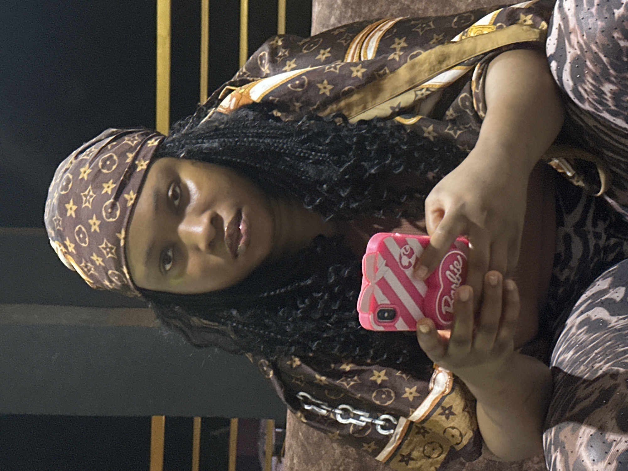 A woman is sitting, looking forward, and holding a cell phone with a pink case. She has dark hair in braids and is wearing a brown headscarf with a star and circle design. She is also wearing a brown jacket with a similar design and gray pants. The photo was taken in the city of Asaba-Ogwashi, Nigeria.
