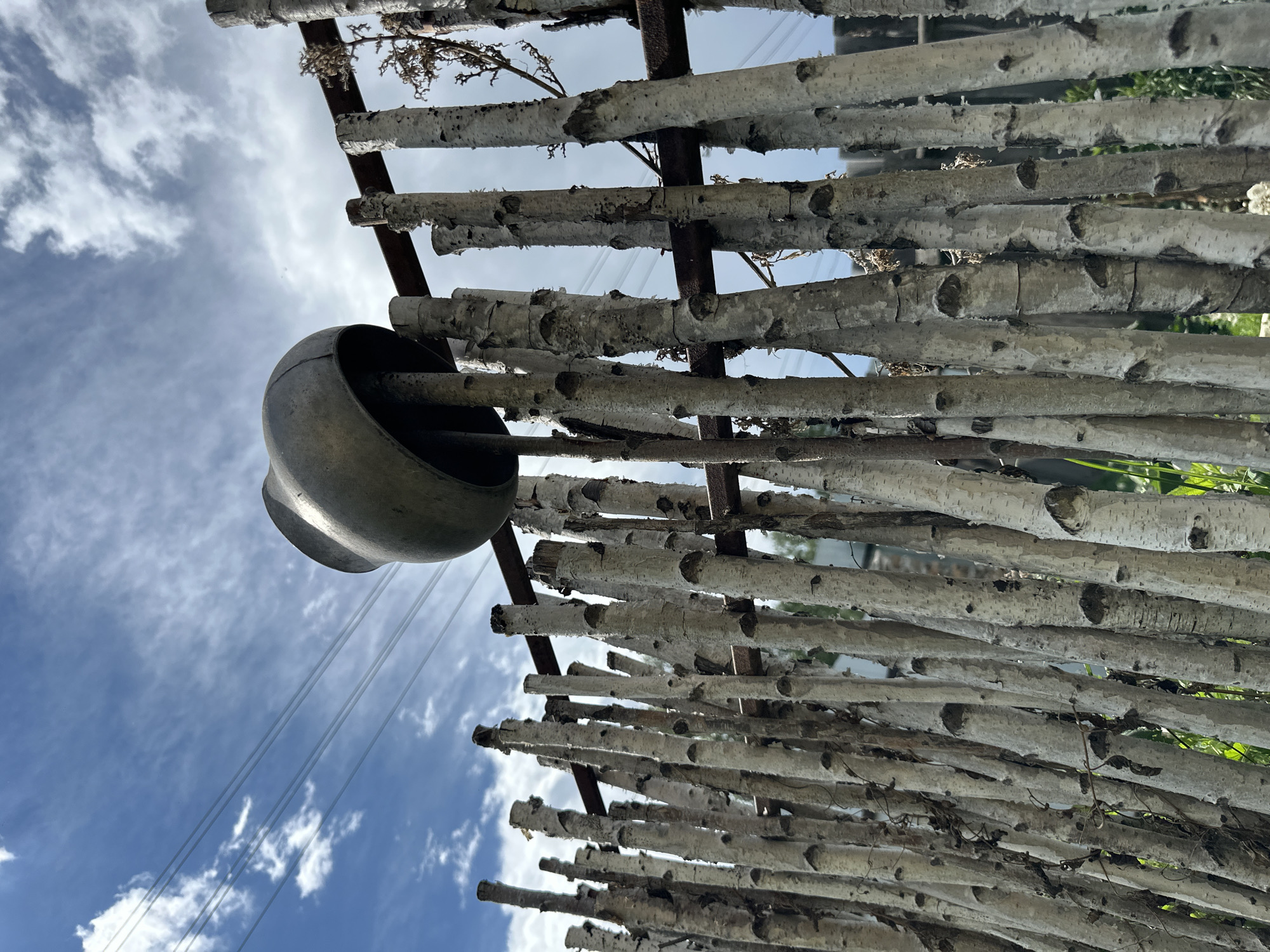 This image captures an outdoor scene in the city of Unknown, Unknown, featuring a fence constructed from vertically oriented, light-colored tree logs, likely birch, exhibiting characteristic peeling bark and dark knots. The logs are secured by two horizontal, rusted metal bars. Perched inverted atop one of the central logs is a dark, metallic, rounded pot. The background consists of a bright blue sky with scattered white clouds, ranging from wispy cirrus to more defined cumulus formations. Several thin, parallel power lines are visible traversing the upper left and central portions of the sky. The scene is brightly illuminated by natural daylight. No people or animals are present.
