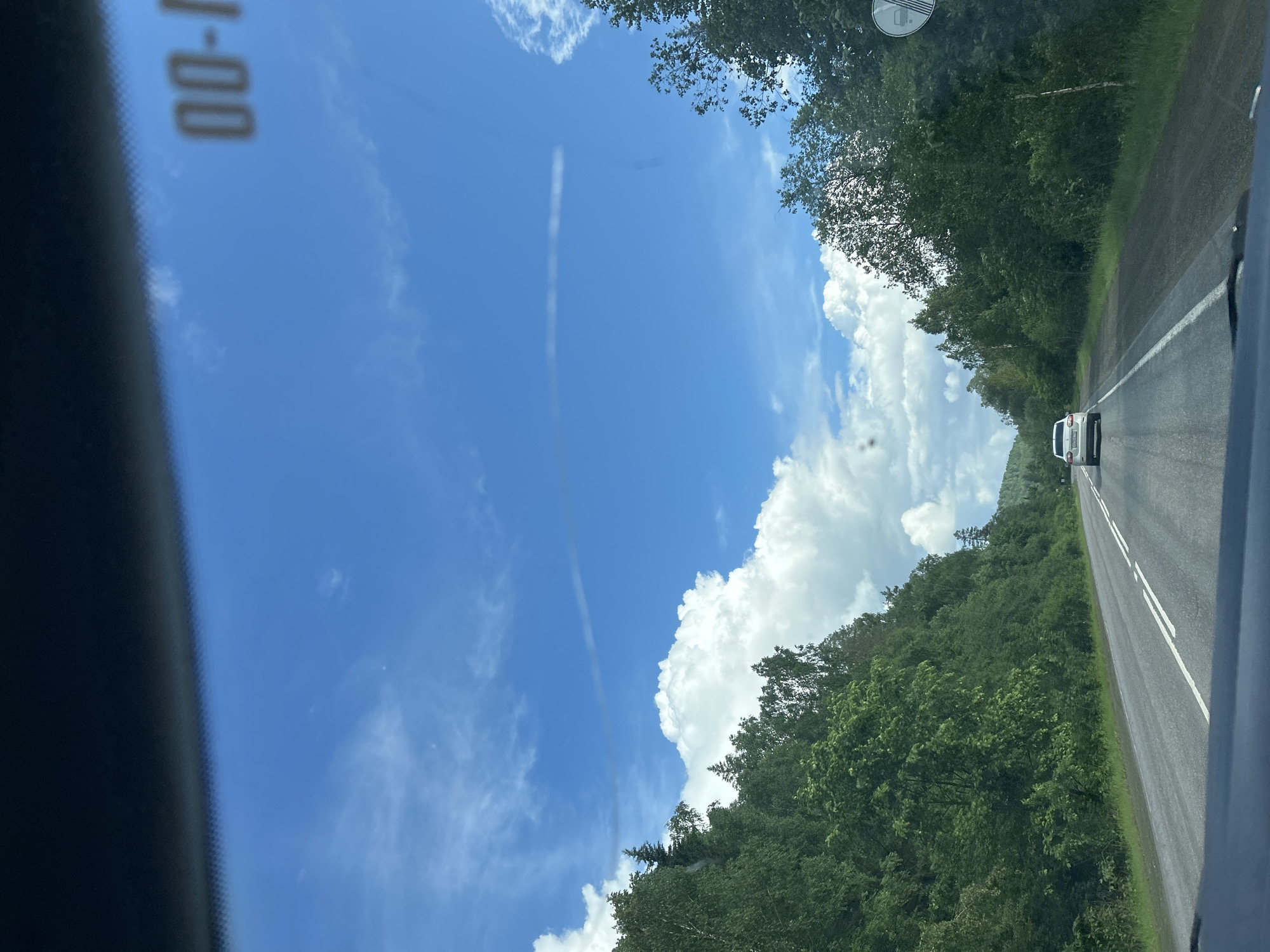 The image captures a view from inside a vehicle looking out at a two-lane asphalt road cutting through a lush, green forest. The sky above is a vibrant blue, adorned with fluffy white cumulus clouds and a faint, elongated cloud streak. Trees line both sides of the road, creating a tunnel-like effect. The road itself has white dashed lines marking the lanes, and a white car is visible ahead, traveling in the same direction. The scene appears to be set in a rural or natural environment, possibly on a highway or a main road through a forested area. There are no visible people or specific landmarks that would definitively place this scene in Усмангали, Russia, though the environment is consistent with many rural areas in Russia.