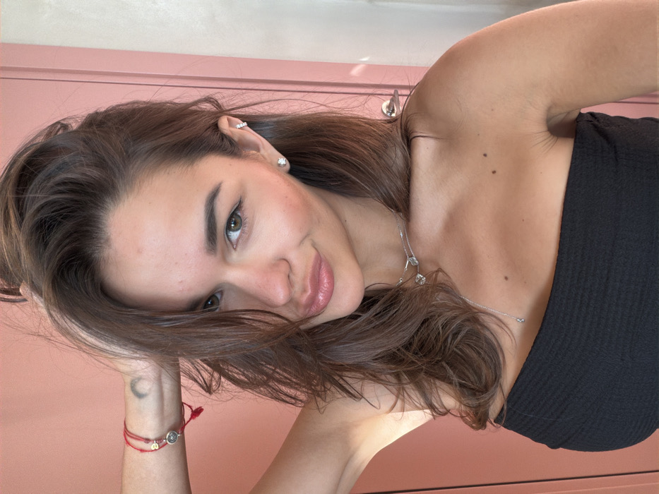 A close-up, chest-up portrait features a female individual with fair skin, dark brown hair, and light brown eyes, looking directly into the camera with a slight smile and pursed lips. Her hair is voluminous, styled to partially obscure her forehead and fall over her shoulders. She wears a black, strapless, textured top. Her right arm is raised, with her hand positioned behind her head, lifting some of her hair.
Accessories include:
- A red string bracelet with a small circular charm and a smaller red bead on her right wrist. A faint crescent moon tattoo is visible on her inner right wrist.
- Two silver-colored necklaces: a choker style with small, clear crystal-like pendants, and a longer, delicate chain with a single small pendant extending down her chest.
- A small, clear stud earring in her left earlobe and a silver-colored multi-segment hoop earring higher on her left helix.
The background is out of focus, depicting an indoor setting. It primarily features a solid light coral-colored surface, likely a wall. To the right, a vertical section of a light-colored door or panel with a silver-colored handle is partially visible. No specific identifying markers are present within the image to forensically determine a precise location in Tel-Aviv, Israel.