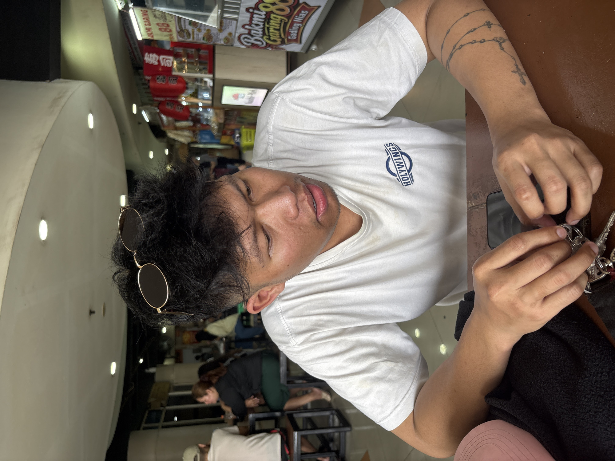 A young man with dark hair and sunglasses perched on his head sits at a wooden table, looking down and to his right. He wears a white t-shirt with the "HOLY WINGS" logo visible on the front. His right hand is in the foreground, manipulating a set of keys. A partial view of a pink hat and a black garment is visible on the left side of the table. In the background, a food court is apparent, with red lanterns and various food stalls. One stall displays a sign for "Bakmi Garing 88" in Indonesian. The ceiling of the food court has circular lighting fixtures. The overall setting appears to be a casual dining area, likely within a shopping mall or market in North Jakarta, Indonesia.