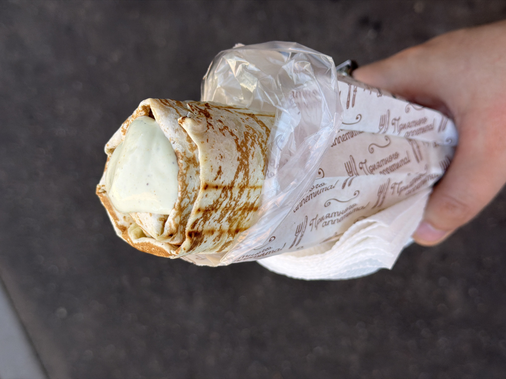 A close-up, eye-level shot shows a hand holding a shawarma wrapped in paper and plastic. The shawarma is topped with a dollop of white sauce. The paper wrap is printed with a repeating pattern of cutlery and text that appears to be in Cyrillic script, possibly indicating a food establishment in Moscow, Russia. The background is a dark, textured surface, likely a sidewalk or road. The lighting is natural, suggesting an outdoor setting.