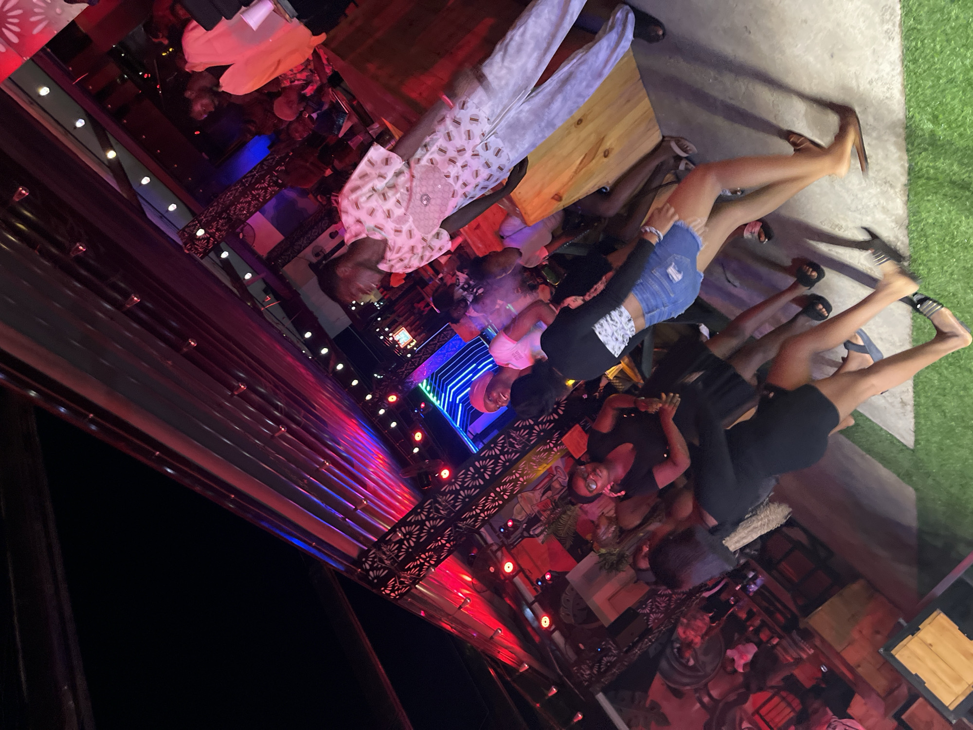 The image depicts a lively nighttime scene at an outdoor venue in Howayi, Nigeria. A group of individuals, predominantly young adults, are engaged in what appears to be social activity, possibly dancing or conversing. The atmosphere is illuminated by colorful lights, with prominent red and blue hues reflecting off the corrugated metal ceiling and structural elements. Several people are visible, with some in the foreground and others in the background. One person in the foreground is wearing a black dress and denim shorts, while another is dressed in a white patterned shirt and white pants. The ground is a mix of concrete and artificial turf. The setting suggests a bar, club, or entertainment space.