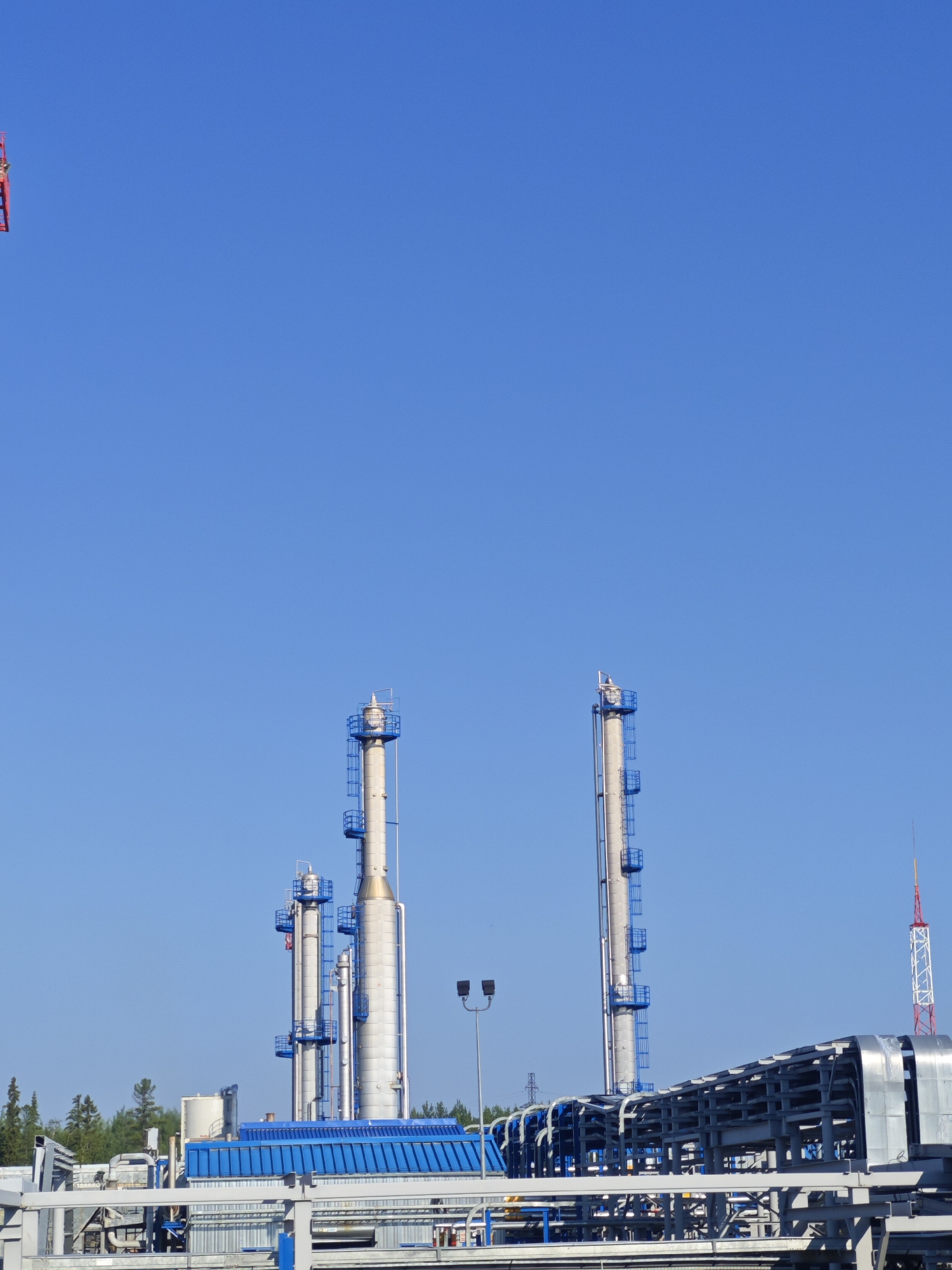 An outdoor, eye-level, medium shot captures a clear blue sky above an industrial facility. The facility features several tall, cylindrical smokestacks with blue platforms and ladders. In the foreground, there is a complex network of metal pipes and structures, some of which are covered with a blue roof. To the far right, a red and white striped tower with a pointed top stands against the sky. Trees are visible in the background, suggesting a semi-rural or industrial park setting. A small, red object with some lettering is visible in the upper left corner, possibly a flag or banner on a crane. No people are visible in the image.