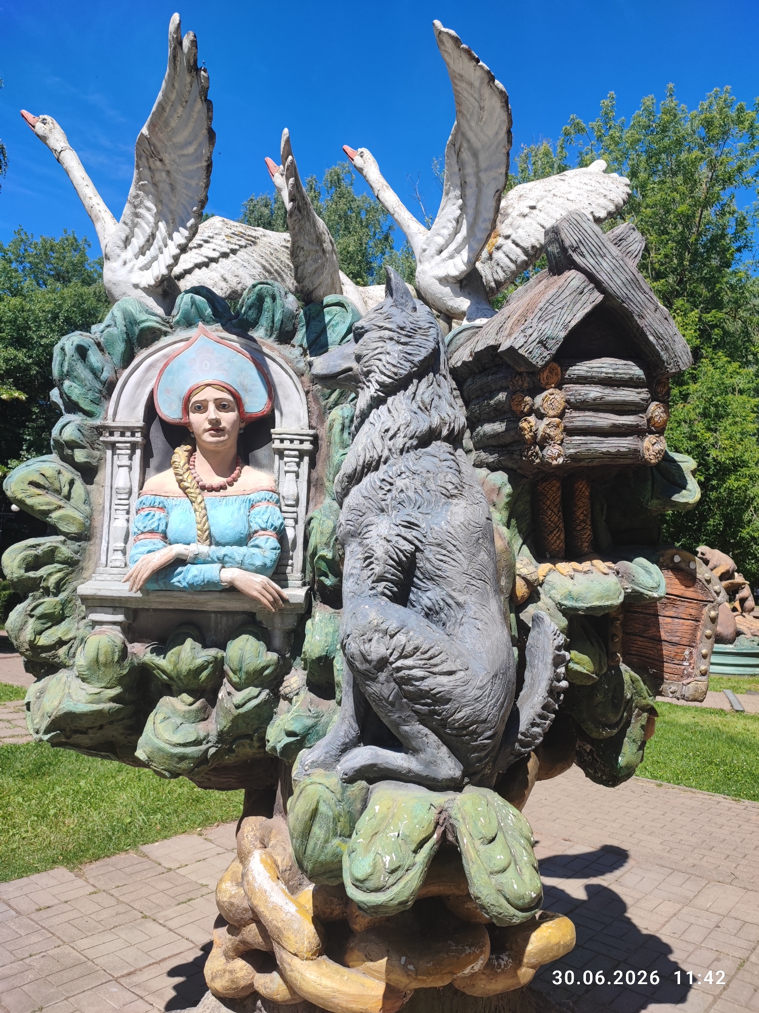 The image shows a colorful outdoor sculpture in Lytkarino, Russia, on a sunny day. The sculpture depicts a scene from a fairy tale and features several characters and elements. In the center, a young woman with blonde braided hair and wearing a blue dress and a traditional Russian headdress is depicted inside an arched frame. To her right, a large, grey wolf statue is seated, looking towards the woman. To the left of the woman, there are three white swan statues with their wings outstretched, their beaks pointing upwards. Adjacent to the wolf and behind the woman is a small, rustic log cabin structure. The entire sculpture is mounted on a base decorated with green, leaf-like shapes and yellow, twisted forms resembling roots or vines. The ground around the sculpture is paved with light-colored rectangular bricks, and a patch of green grass is visible to the left. Lush green trees form the background. The bottom right corner of the image includes a timestamp: "30.06.2026 11:42".