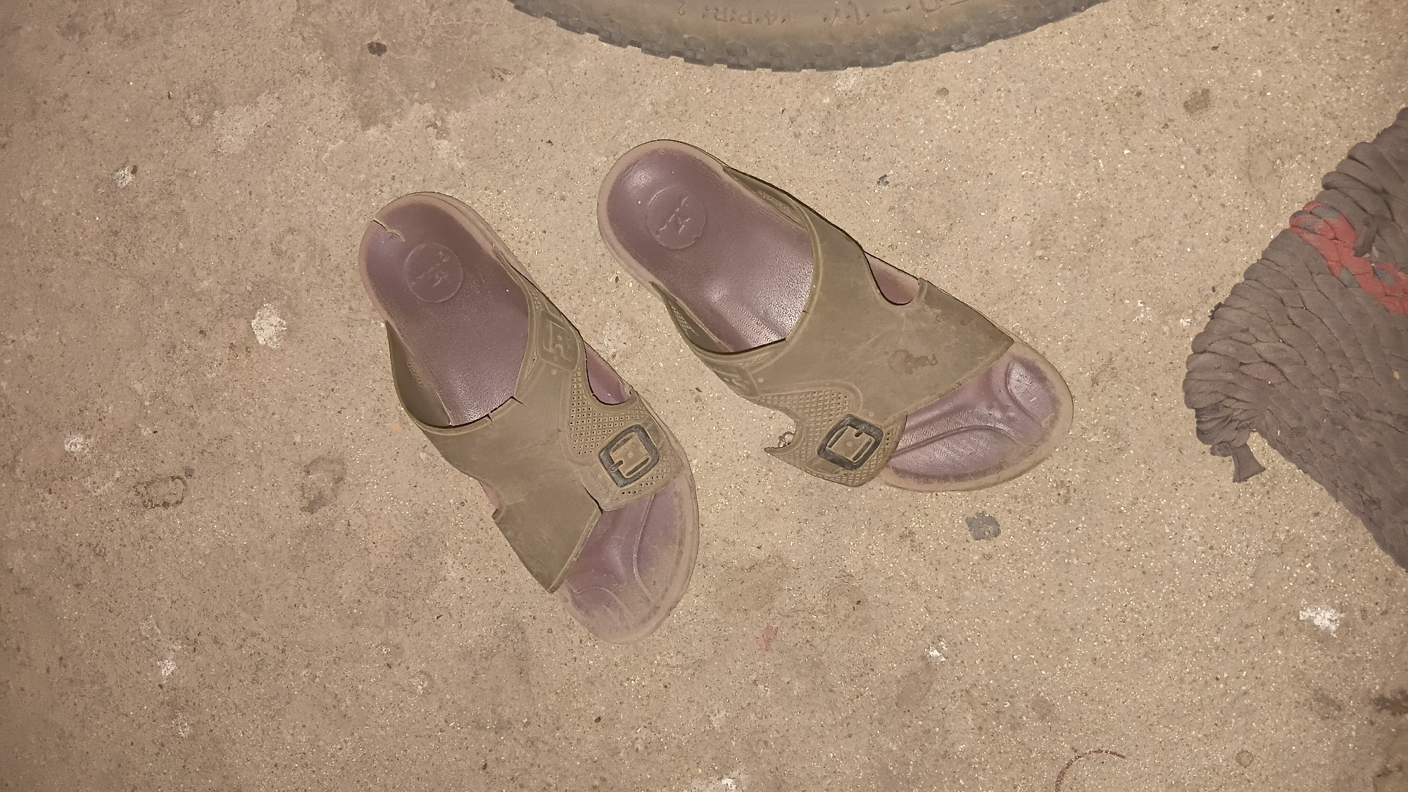 Two dusty, brown slide sandals are lying on a concrete surface. The sandals are positioned at an angle, one slightly above and to the left of the other. The concrete surface appears to be outdoors, possibly a sidewalk or patio, with some small white specks and darker marks scattered across it. In the lower right corner of the image, a portion of a tire is visible, suggesting the presence of a vehicle nearby. At the bottom center, a crumpled, dark gray cloth with a red stripe is also visible. There is no indication of people or any specific action taking place, other than the placement of the sandals on the ground. The image does not provide enough information to determine its specific location within Prabumulih, Indonesia.