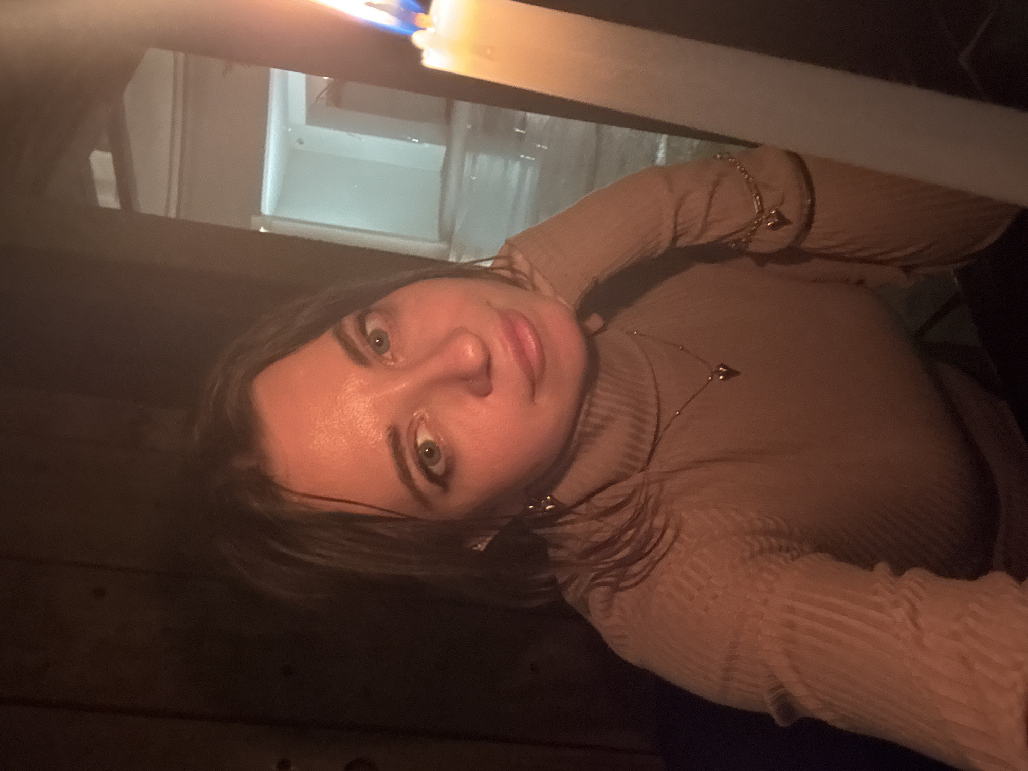 A woman is holding a lit candle. The location may be Moscow, Russia. She wears a tan turtleneck sweater and gold jewelry. The background is dark and blurry, possibly showing a doorway or window with a glimpse of a brightly lit interior. The photo appears to be taken at night.