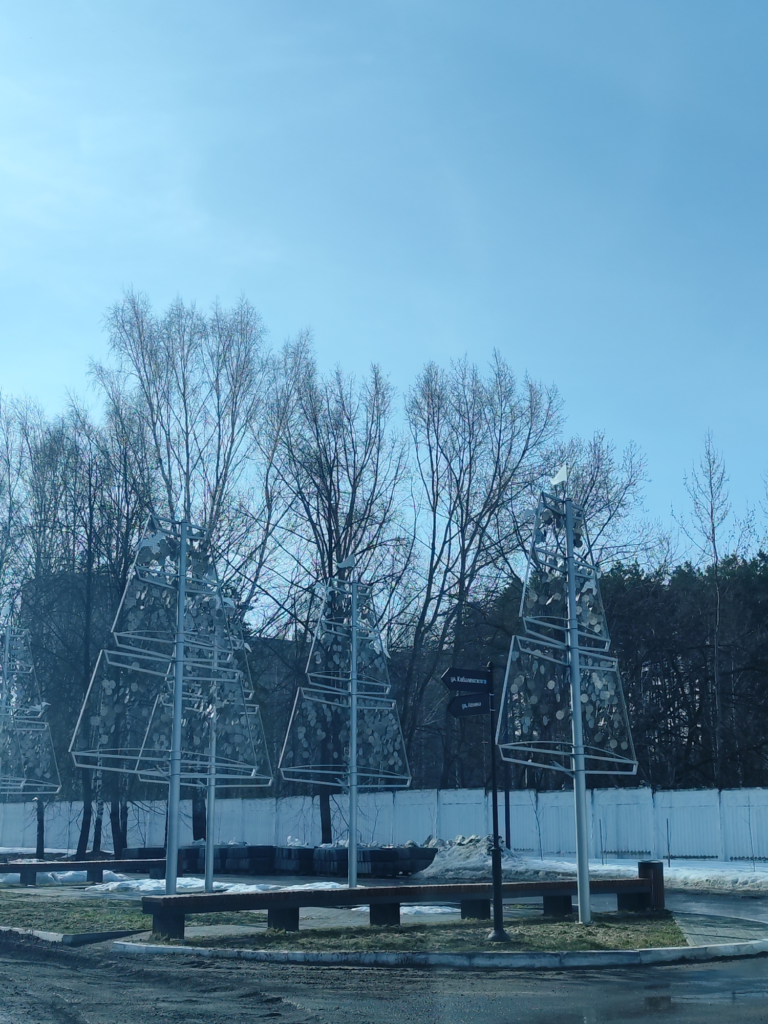 The image depicts an outdoor scene in Chaikovsky, Russia. In the foreground, there are several decorative metal structures shaped like stylized Christmas trees, arranged in a row. These structures are placed on small circular patches of grass. Benches are situated in front of these structures. Beyond the decorative trees, there is a line of bare deciduous trees without leaves, followed by a coniferous forest in the background. A white fence runs along the edge of the wooded area. A street sign is visible near one of the metal structures, indicating the direction to "Ul. Kabalevskogo" and another street, "Ul. Lenina". Patches of snow are visible along the side of the fence. The foreground is a paved area, likely a parking lot or street, with some damp spots. The sky is blue and relatively clear. The overall scene suggests a park or recreational area in a city during the late winter or early spring season. There are no people visible in the image.