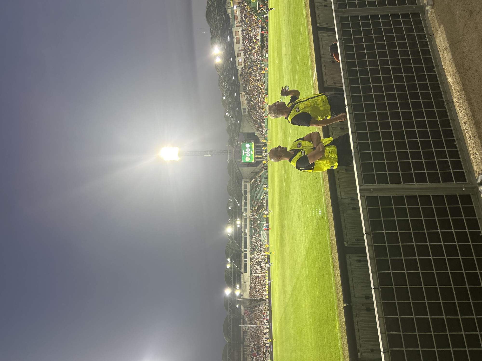 The image depicts a football match at the stadium in Žilina, Slovakia. Two security personnel in yellow vests stand in the foreground, facing away from the camera, with one drinking from a bottle. Behind them, the green football field is illuminated by stadium lights, and players in yellow and green uniforms are visible. The stands, filled with spectators, are in the background, under a cloudy sky. A large scoreboard shows a score of 0:0. The stadium appears to be modern and well-maintained.