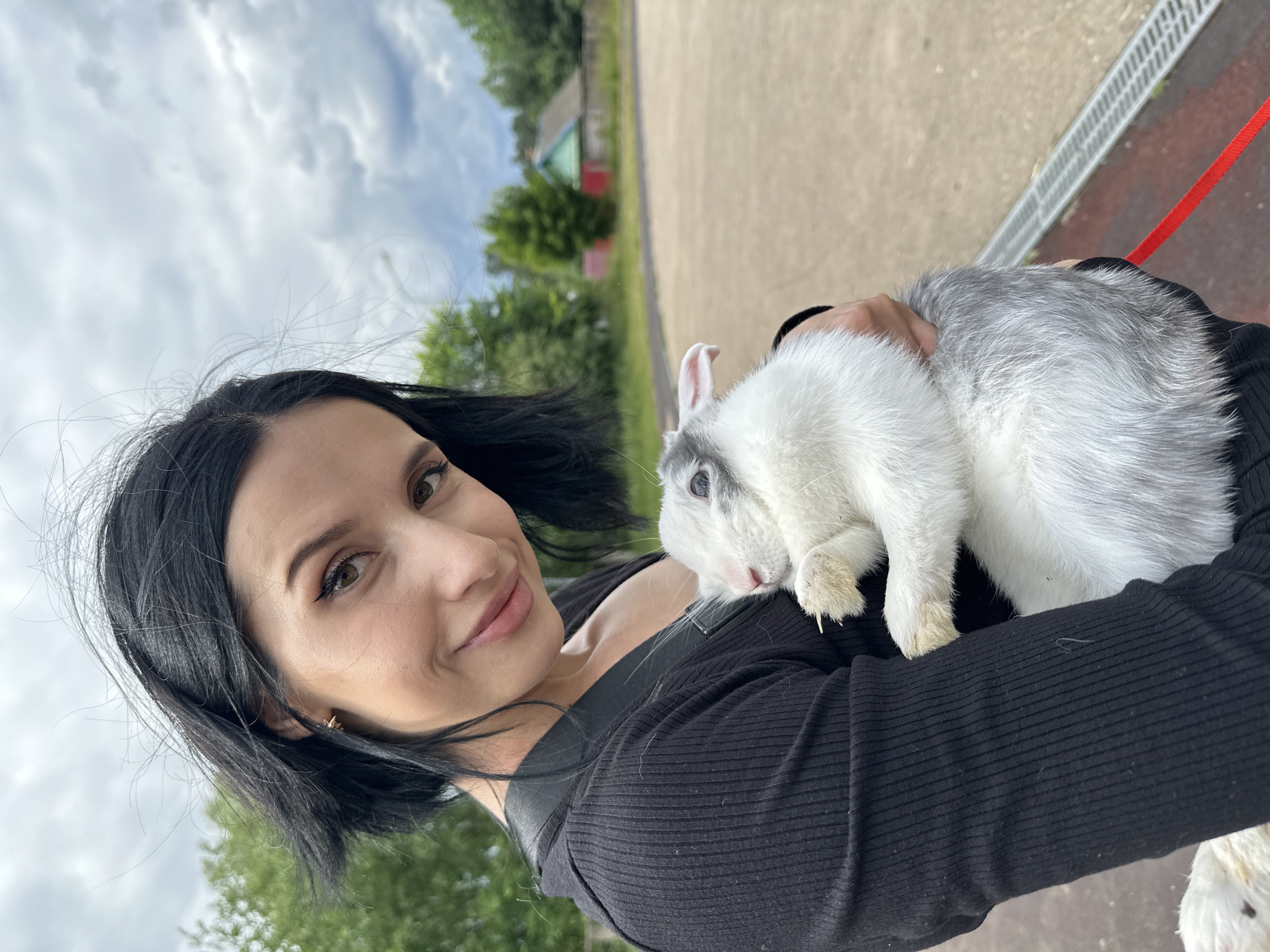 In Chaikovsky, Russia, a woman with black hair and dark eyeliner smiles at the camera while holding a white and gray rabbit. She is wearing a black ribbed top. The rabbit is nestled in her arms, with its head resting on her chest. The background shows a cloudy sky, green trees, a small rural house with a red and green facade, and a paved area.