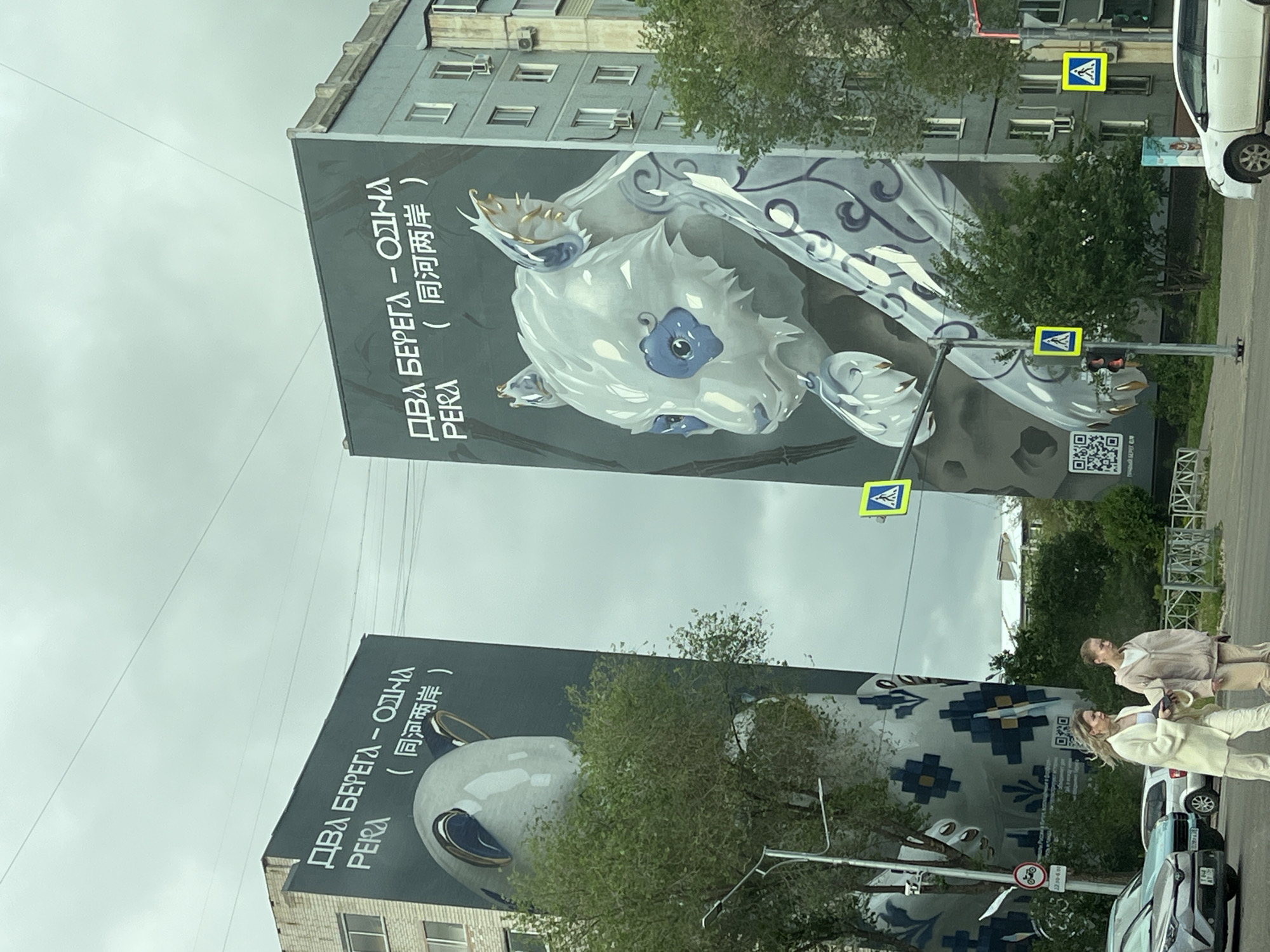 The image shows two tall buildings in what appears to be an urban environment, likely a city in an area where both Russian and Chinese languages are understood. Large murals cover the sides of these buildings. The murals feature an artistic design of a whale-like creature with intricate patterns. The Russian text "ДВА БЕРЕГА - ОДНА РЕКА" and its Chinese translation "同河两岸" (Tóng hé liǎng'àn) are displayed on both murals, which seems to be the title of the mural. There is a QR code on the upper mural.
In the foreground, there are two women dressed in light-colored clothing walking on the sidewalk, alongside parked cars. Street signs indicating a pedestrian crossing are visible, as well as traffic lights. The sky is overcast, suggesting a cloudy day. The location is described as in the city of Unknown, Unknown.