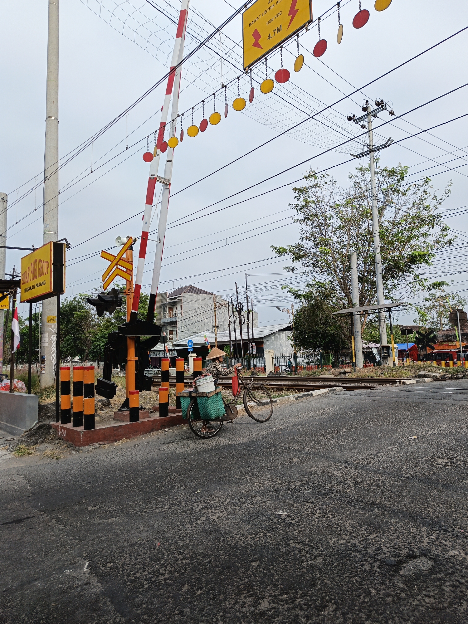 The image depicts a street-level view of a railway crossing in Klaten, Indonesia. A red and white striped railway crossing gate is lowered, indicating an approaching train. The gate is supported by a tall concrete pole on the left, and a shorter pole with black and orange striped bollards on the right. Above the crossing, numerous overhead wires are visible, typical of an electrified railway line, with a yellow sign warning of "KAWAT LISTRIK ALIR 1500 VDC 4.7M" (Live Electrical Wire).
In the foreground, a bicycle with a woven green basket on the back is parked. A person wearing a conical hat, likely a farmer or street vendor, is seated on the bicycle, facing away from the camera. The bicycle is laden with what appear to be containers or bags.
The road surface is asphalt, showing signs of wear and tear. In the background, there are buildings, including a two-story house and smaller structures, as well as trees and vegetation. The sky is overcast, suggesting cloudy weather. The overall scene conveys a sense of daily life in a semi-urban or rural area of Klaten, with the railway crossing being a prominent feature.