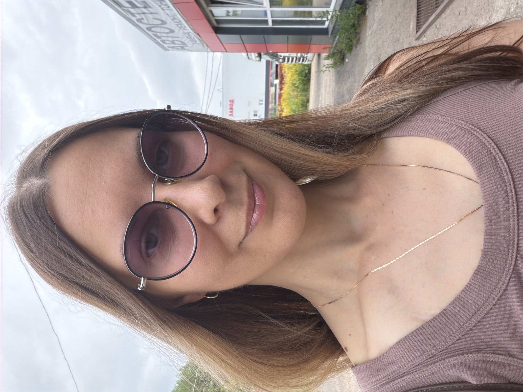 A medium shot photograph shows a woman with long, blonde hair and sunglasses. She is wearing a mauve tank top and a gold necklace. She is smiling faintly. Behind her, a white building with "ЗАВОД" (factory) written on it is visible. To the right, a red and grey building with an "ABTOCTE" sign (likely a car service) is also present. The location is the city of Yagul, Russia. The sky is overcast.