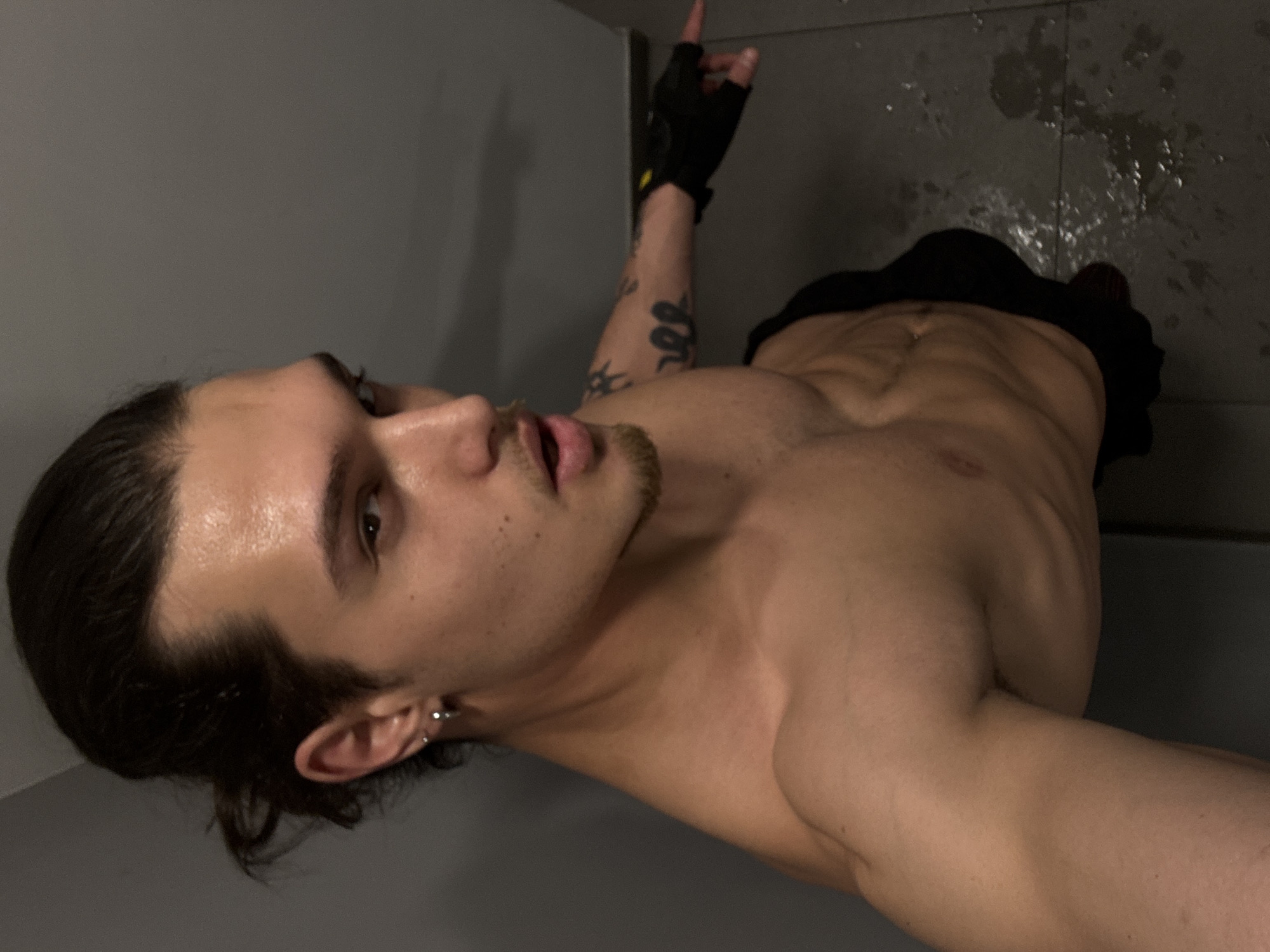 A young adult male, shirtless, is prominently featured in a medium close-up, angled from below and to the side. He possesses a muscular physique, with visible abdominal muscles and defined shoulders and arms. His dark hair is pulled back, with some strands framing his face, and he has a small goatee. He wears a silver hoop earring in his left ear. His skin appears slightly sweaty or glistening.
His left arm is extended upwards and angled to the right, with his index finger pointing towards the top right corner of the frame. He wears a black fingerless glove on this hand. Distinct tattoos are visible on his left arm: a snake design and a sun or starburst pattern. His mouth is slightly open, and his expression is focused and intense as he looks towards the right. He is wearing low-slung black shorts or underwear, visible around his waist.
The background consists of plain gray walls on the left and a darker gray tiled floor on the right. The floor shows visible water splashes or marks, suggesting a wet environment, possibly a shower area, bathroom, or locker room. The lighting is relatively dim and even, consistent with an indoor setting. There is no visible text in the image. The location context provided for this media file is Sofia, Bulgaria.