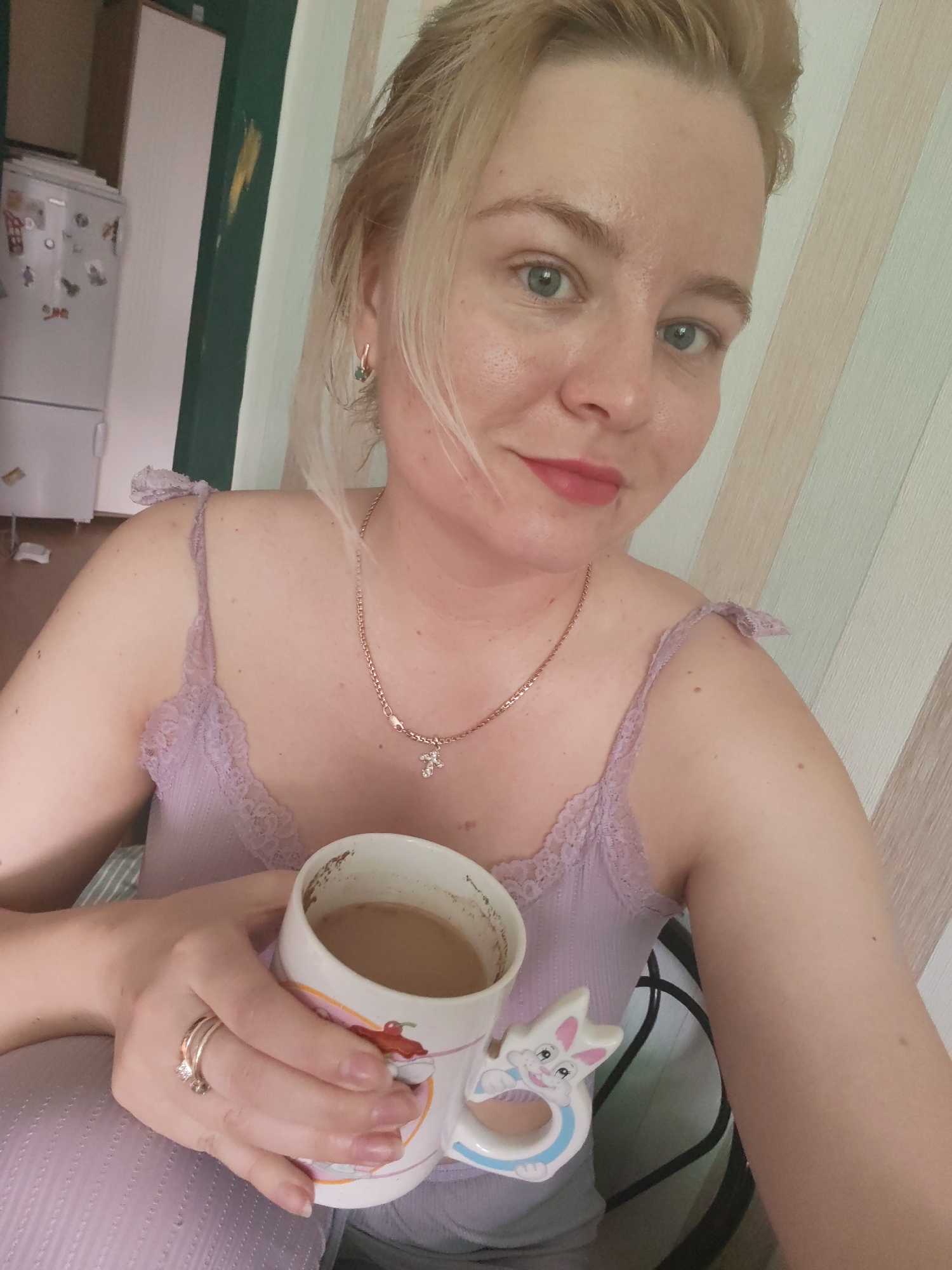 A woman is holding a mug filled with coffee or a similar beverage. She wears a light purple camisole with lace details, a gold necklace, and rings on her hand. The mug has a rabbit-shaped handle and a design on the side. The background shows a striped wall and a glimpse of a room interior with a refrigerator. There is no information or visual evidence indicating that the location is Izhevsk, Russia.