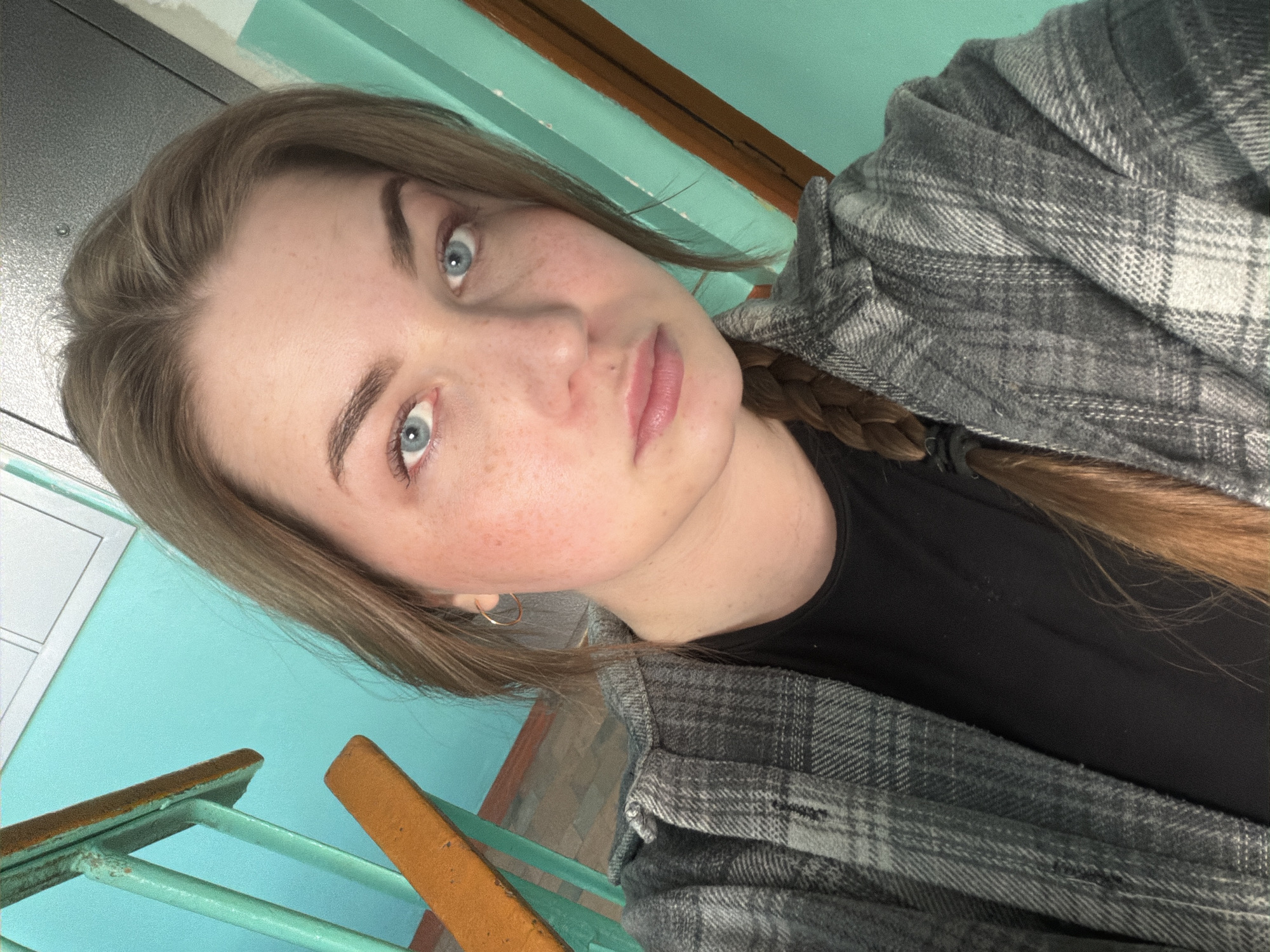 A young woman with light brown hair and blue eyes is shown in a close-up selfie. Her hair is partially braided and she is wearing a gray plaid jacket over a black shirt. She has freckles on her face and is wearing small hoop earrings.
The background consists of a teal wall, a staircase with green railings and brown wooden steps, and a gray door. The location is Chaikovsky, Russia.