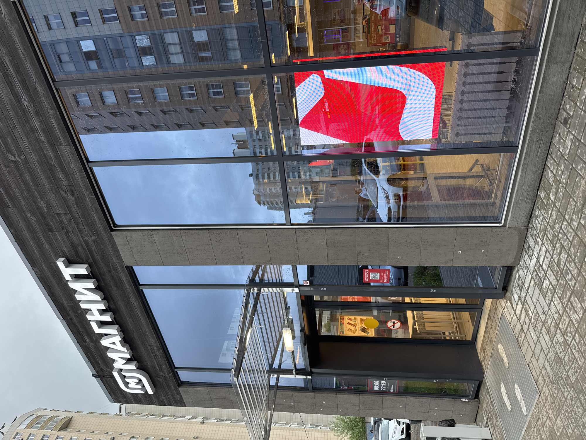 The image depicts the facade of a "Magnit" store, identifiable by the white lettering on a dark-colored, possibly wooden-paneled exterior. The store appears to be situated in an urban setting, suggested by the reflection of multi-story buildings in the store's large windows. The entryway to the store is visible, marked by a metal awning structure. A sign indicating operating hours ("08:00-22:00") is posted near the entrance, along with a yellow and red no smoking sign. The reflection of a large video display inside the store is visible in one of the windows, featuring a red and white graphic. The image is taken at ground level, with wet pavement visible in the foreground. The presence of buildings in the reflection suggests an urban location. Based on the "Magnit" chain and architectural style, it is reasonable to infer the location is in a Russian city, potentially Saint Petersburg given the presence of other European buildings in the reflection of the glass panes. A white car is parked on the street. A rubbish bag is visible near the car.
