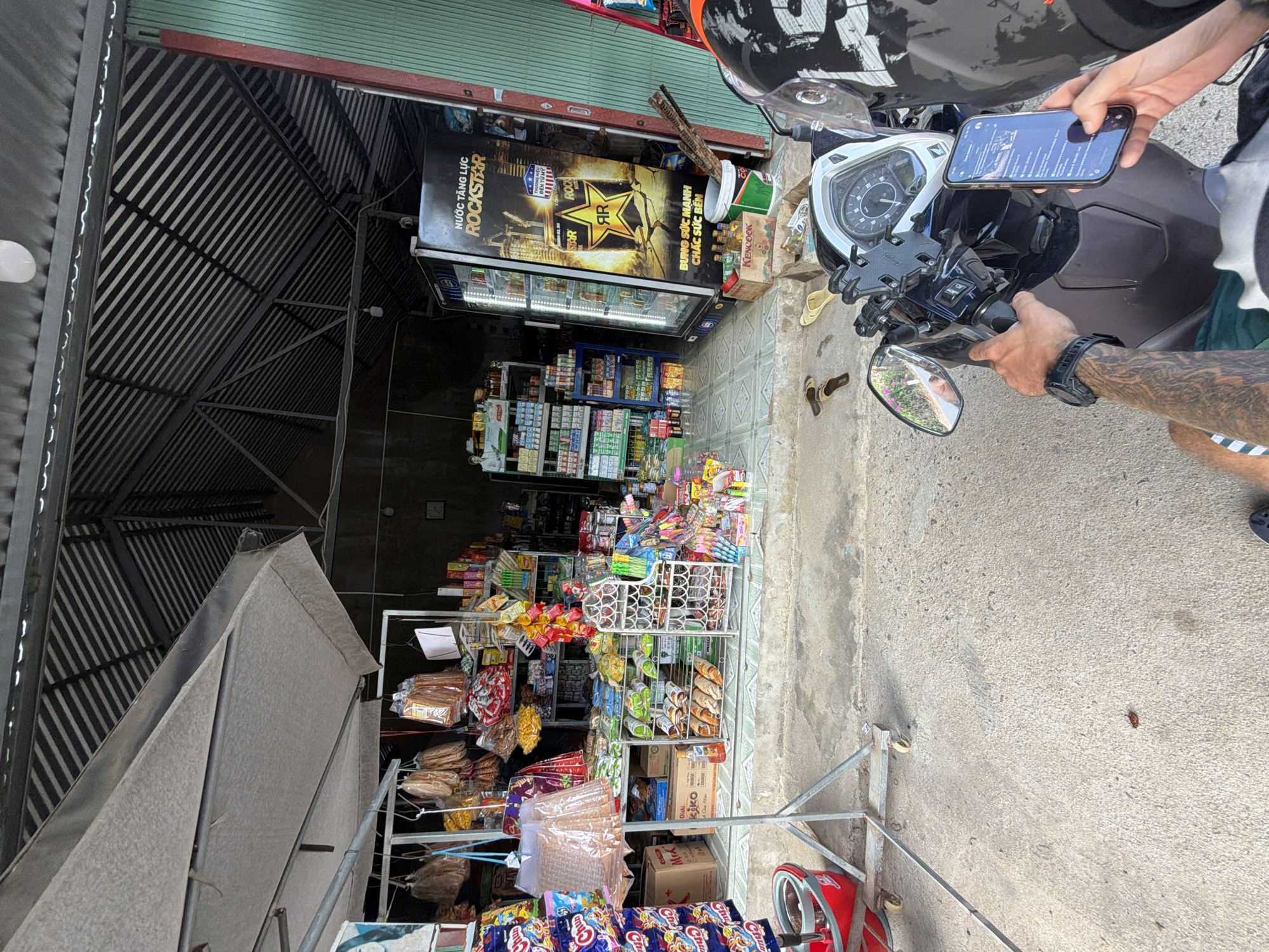 Here's a detailed description of the image:
**Location:** Diên Thọ, Vietnam.
**Overall Scene:** The image shows the storefront of a small convenience store, heavily stocked with a variety of goods. A person is present, partly seen, operating a motorcycle and using a mobile phone.
**Storefront:** The store is densely packed with merchandise. There are multiple racks and shelves displaying snacks, drinks, and various other items. A large refrigerator with "Rockstar" energy drink branding is visible. The store has a corrugated metal roof structure.
**Person:** A person is partially visible, positioned beside a motorcycle. They are holding a mobile phone, apparently using it while on the motorcycle. Part of their arm is visible, showing tattoos.
**Motorcycle:** A motorcycle is present, parked in front of the store. Its dashboard and handlebars are visible, along with a phone mount.
**Environment:** The ground in front of the store is paved.