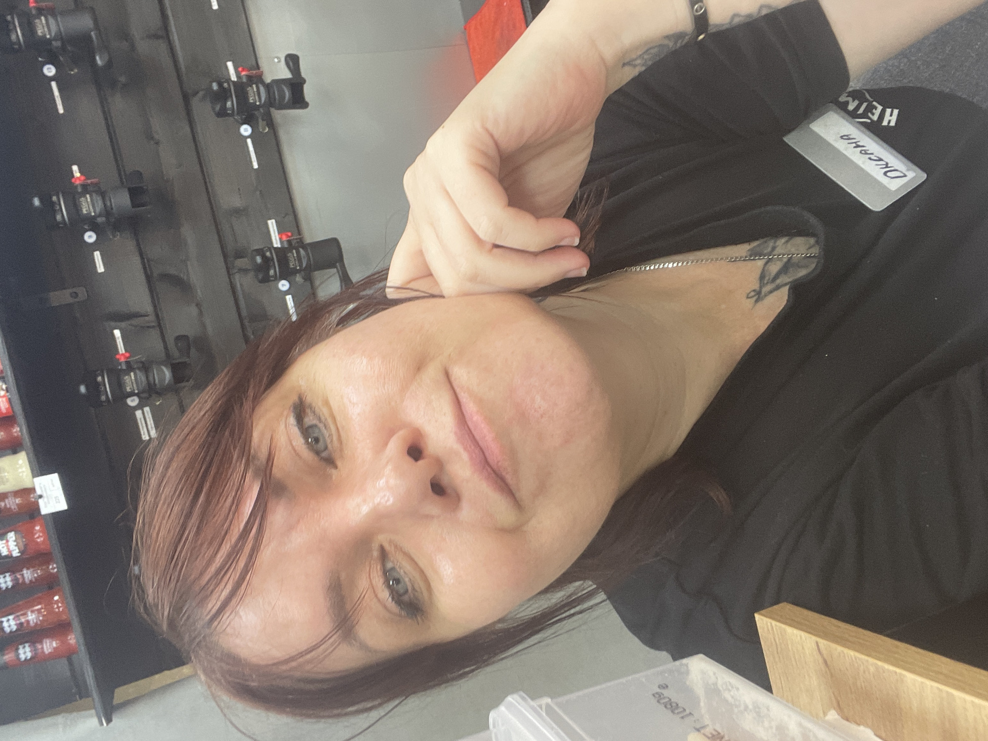 The media file is a portrait of a person, likely a woman, taken in a vertical orientation. She has brown hair and is looking towards the camera with a neutral expression. Her right hand is resting on her cheek, and her left hand is tucked behind her head, partially obscuring her hair. She is wearing a black shirt with a silver necklace and a name tag that reads "ОКСАНА" (Oksana) in black lettering on a white rectangle. There are tattoos visible on her arm and chest.
In the background, to the left, there is a dark wooden shelf with several black objects mounted on it, possibly taps or dispensers. To the far left, on another shelf, are rows of red packaging with white text. To the right of the person, a wooden surface is visible. The overall setting appears to be an indoor establishment, possibly a bar, restaurant, or store.
The lighting suggests it is daytime, and there are no clear indications of the weather. The location context provided is Rusanovo, Russia. The image conveys a casual and personal moment, possibly a selfie.