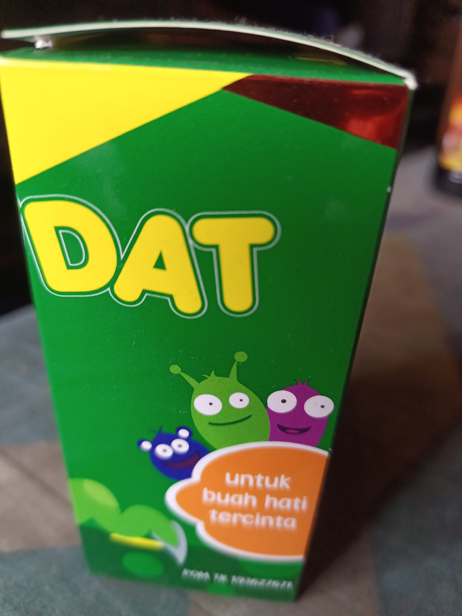 The image shows a close-up of a green box with yellow lettering that reads "DAT". The box has a diagonal yellow and red design on the top left. Below the lettering, there are three cartoon aliens with large eyes and mouths. The alien on the left is blue and has small ears. The alien in the middle is green and has a smiley face. The alien on the right is purple and also has a smiley face. To the right of the aliens, there is an orange cloud shape with white text that reads "untuk buah hati tercinta". There is also some text at the bottom of the box, but it is blurry and illegible. The box is sitting on a surface that appears to be a table or counter. The background is dark and out of focus. There is no information to suggest the location is in South Jakarta, Indonesia.